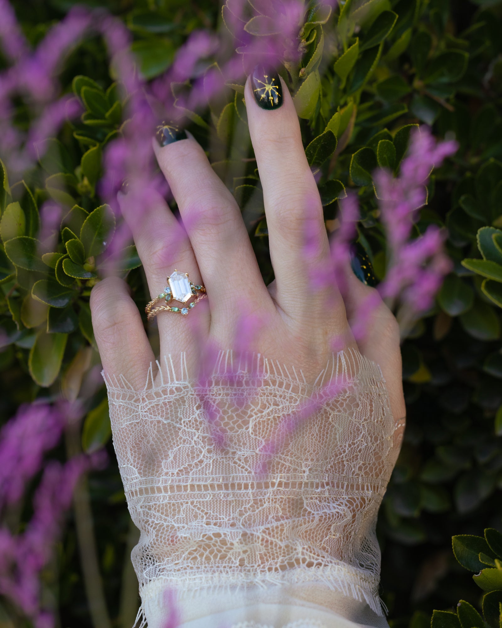 Hand wearing a ring with a moissanite gemstone, surrounded by purple flowers and green leaves.
