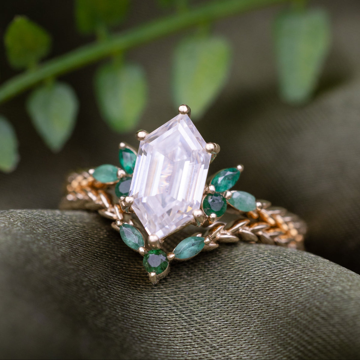 A gold ring with a hexagon Moissanite center stone and emerald accent stones, displayed on a textured olive green surface with blurred green leaves in the background.