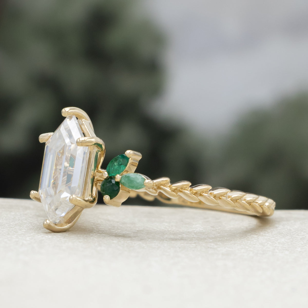 A close-up of a gold ring with a hexagon-cut moissanite and multiple green emerald accents against a blurred natural background.