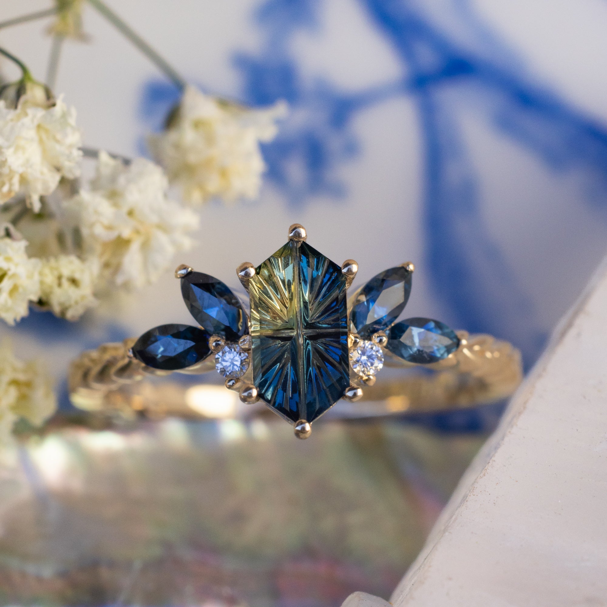 Gold ring with Australian sapphire center stone and diamond accents in front of white flowers