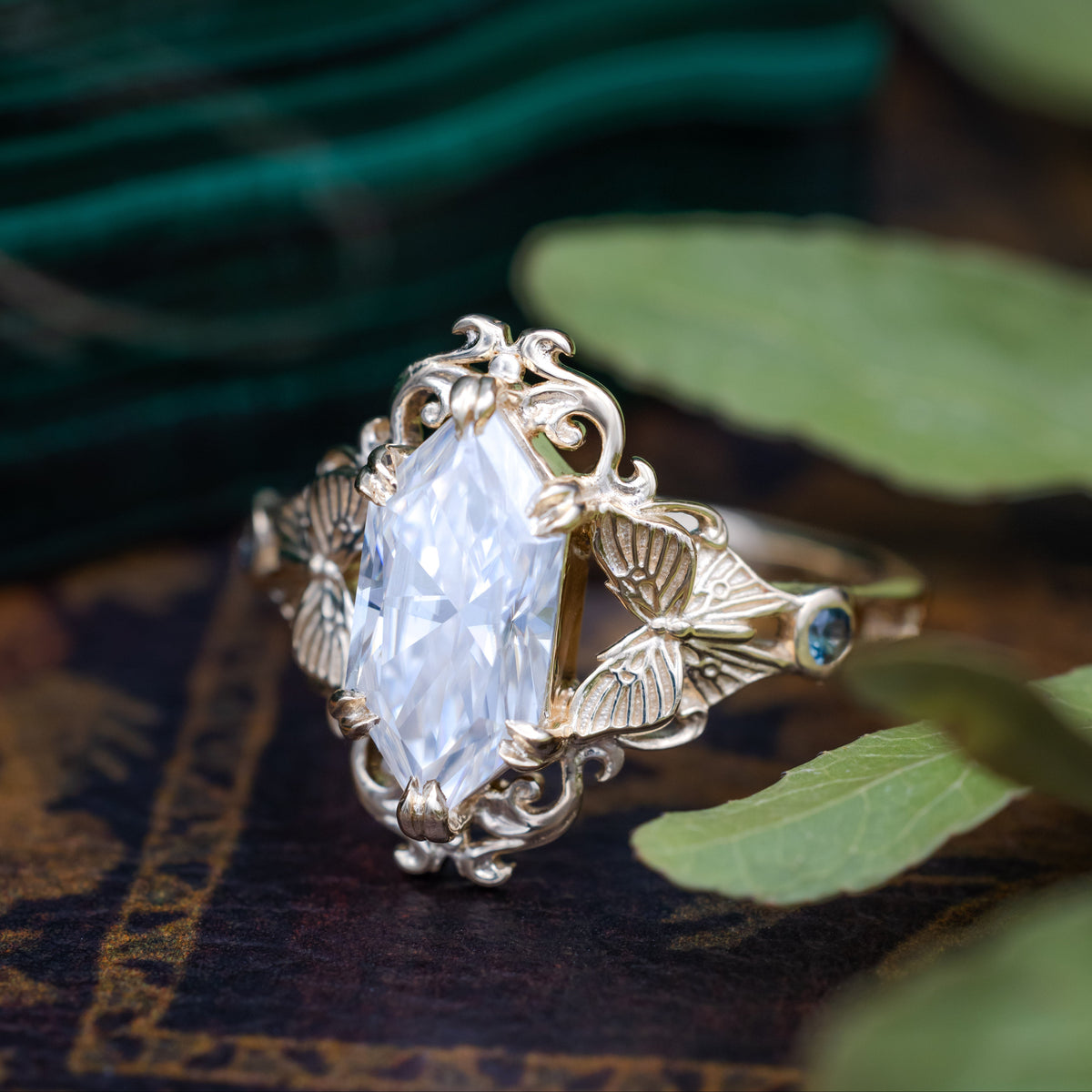 A gold ring with moissanite center stone, moth filigree and sapphire accents against a decorative background