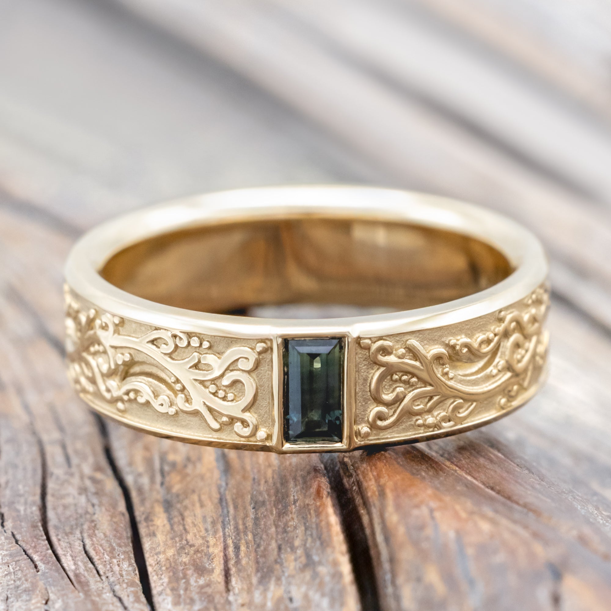 A gold ring with intricate floral engravings and a blue-green sapphire resting on a wooden surface.
