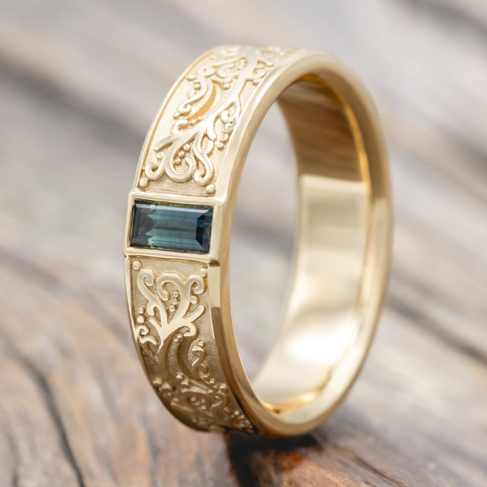 A gold ring with intricate floral engravings and a blue-green sapphire resting on a wooden surface.