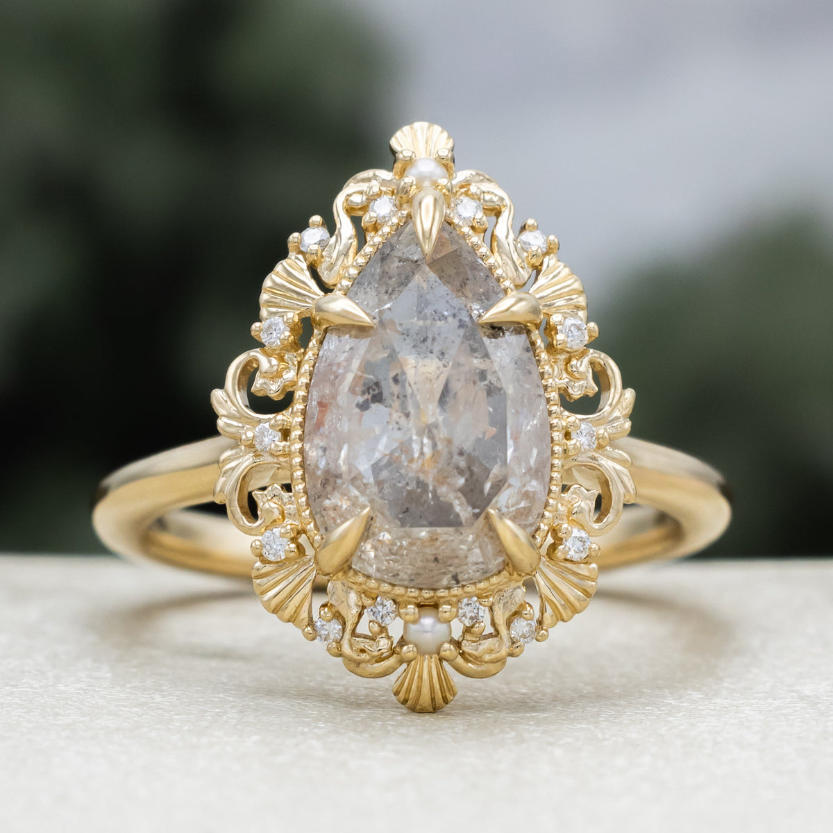 A sculptural gold ring with large pear cut salt & pepper diamond with pearl and diamond accents against a blurred green background