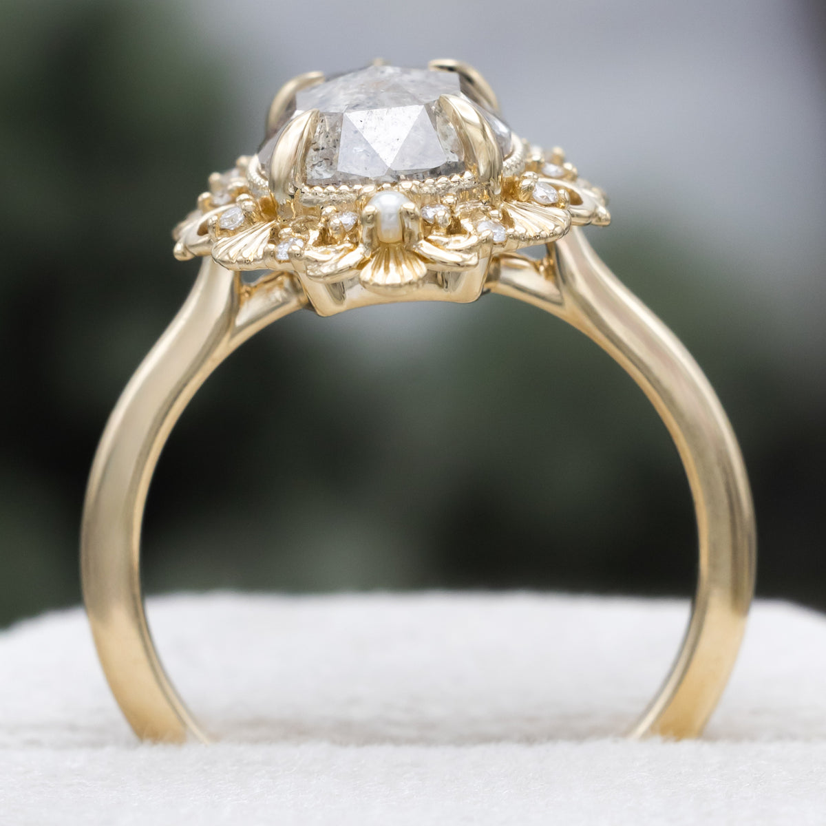 A sculptural gold ring with large pear cut salt & pepper diamond with pearl and diamond accents against a blurred green background