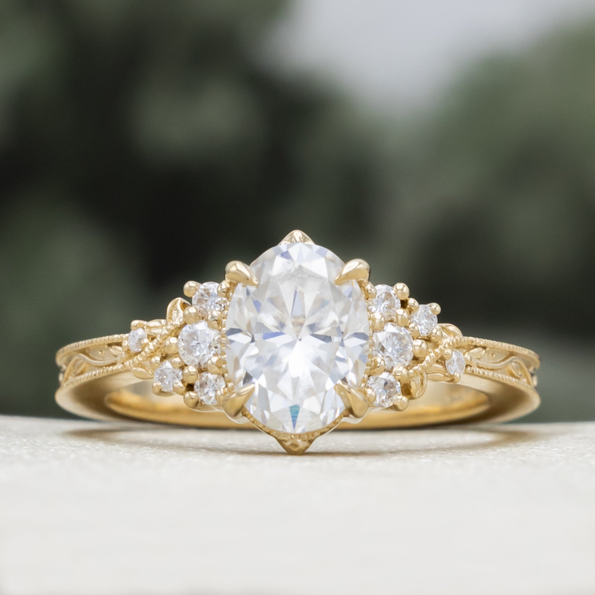 A gold engagement ring with an oval Moissanite and diamond accents, against a blurred green background