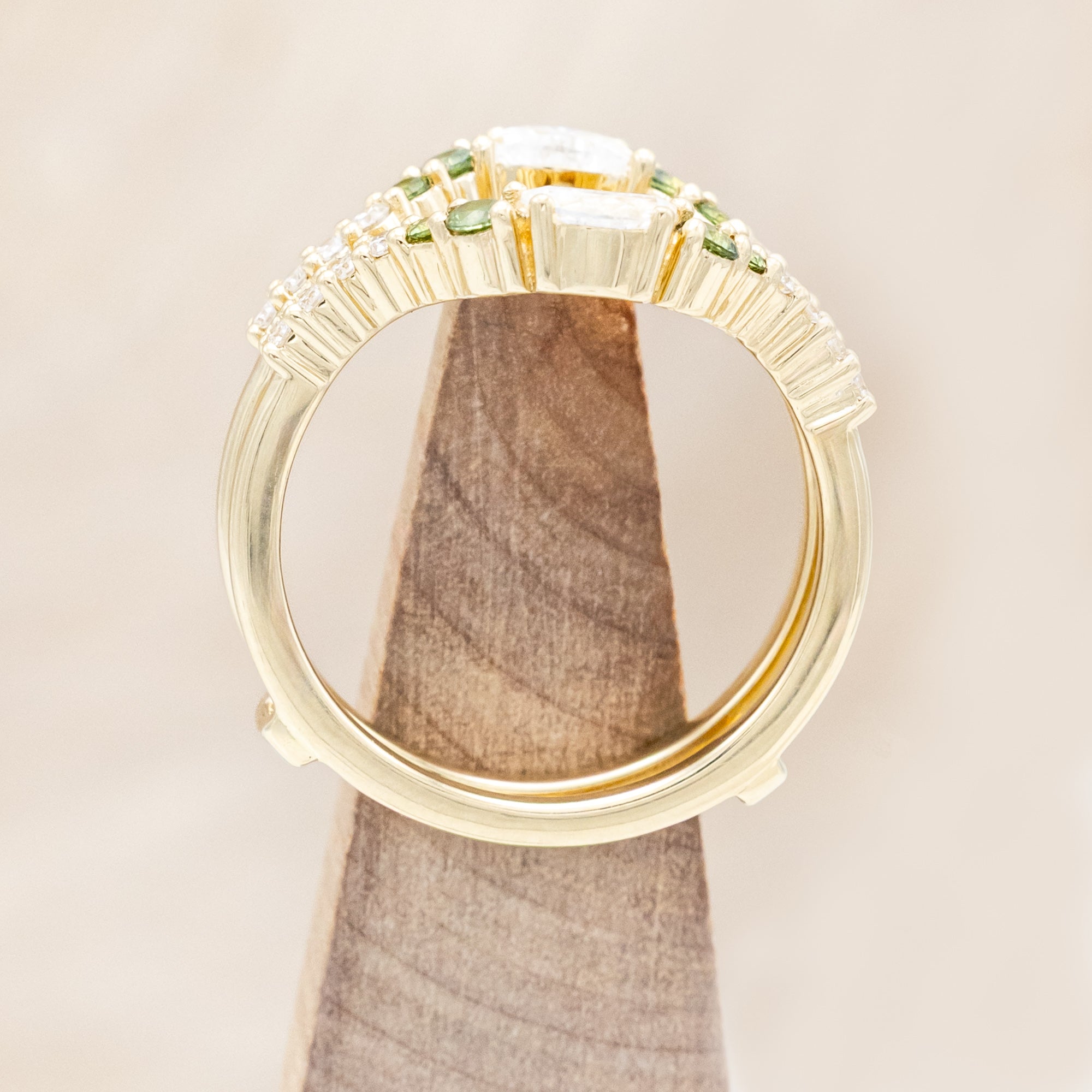 Gold ring with moissanite, green sapphire & diamond accents on a wooden stand against a beige background
