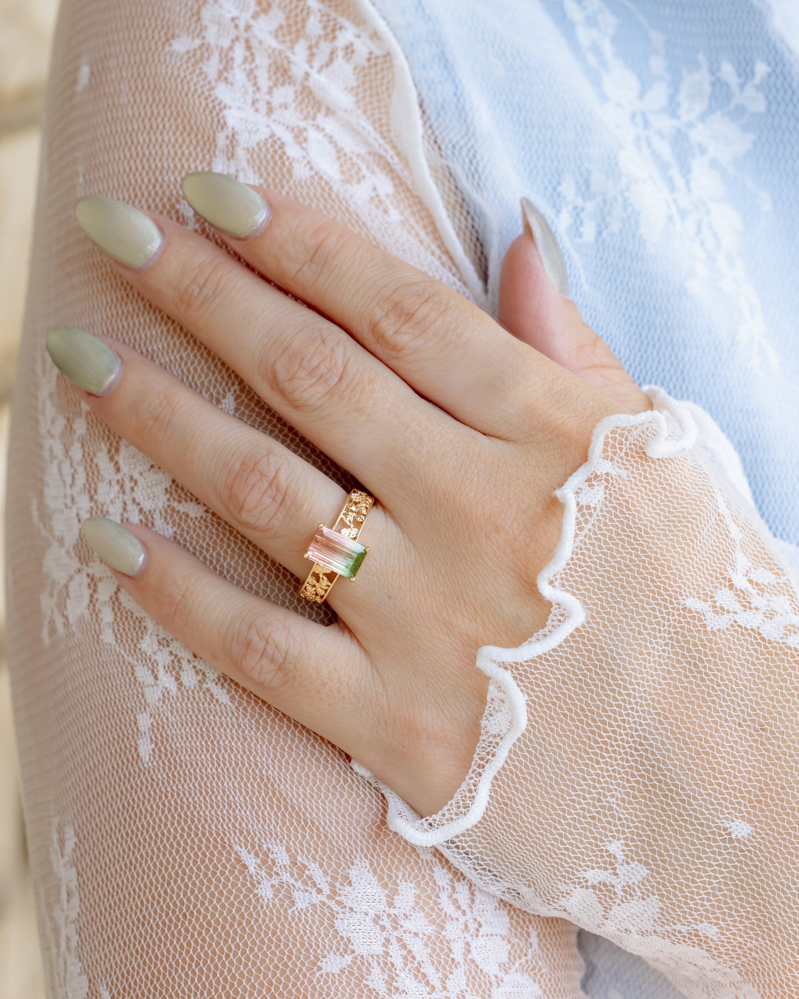 Hand wearing a gold ring with green and pink torumaline gemstones on a lace fabric background