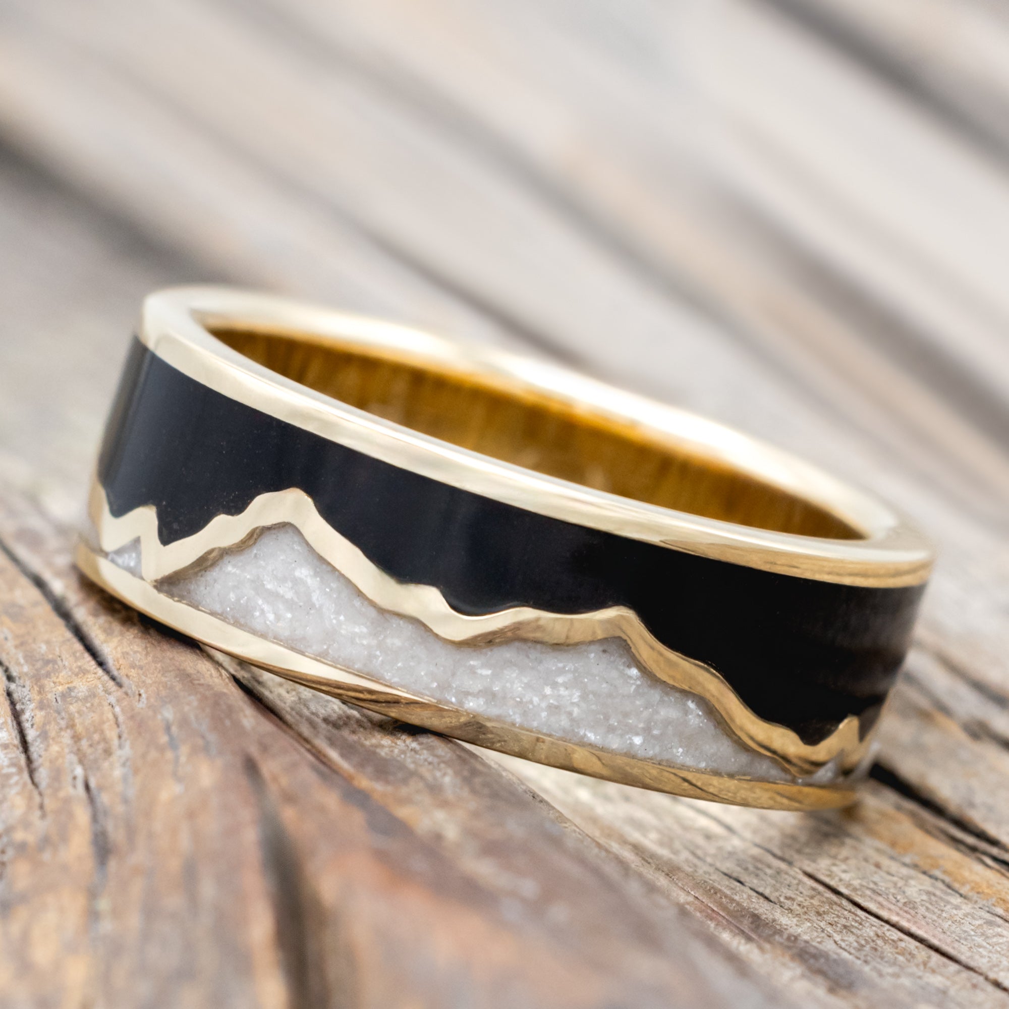 A gold ring with jet stone sky and diamond dust inlays with gold mountains between the two on a wooden surface