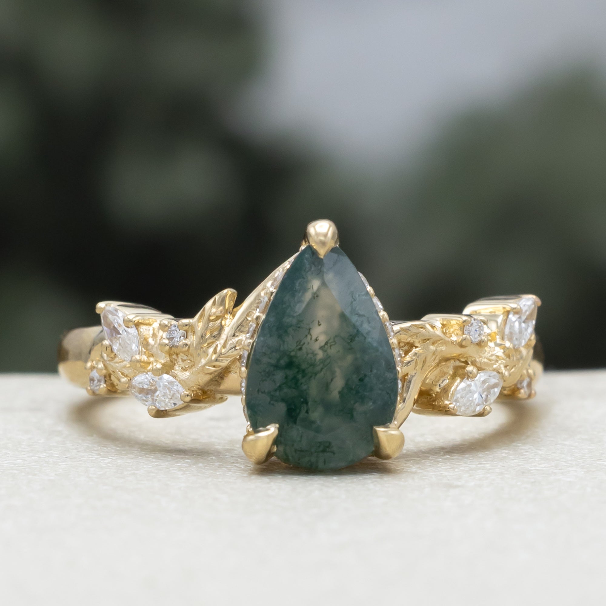 A gold ring featuring a pear cut moss agate flanked by delicate gold leaf-like branches with diamond accents, set against a blurred dark green background.