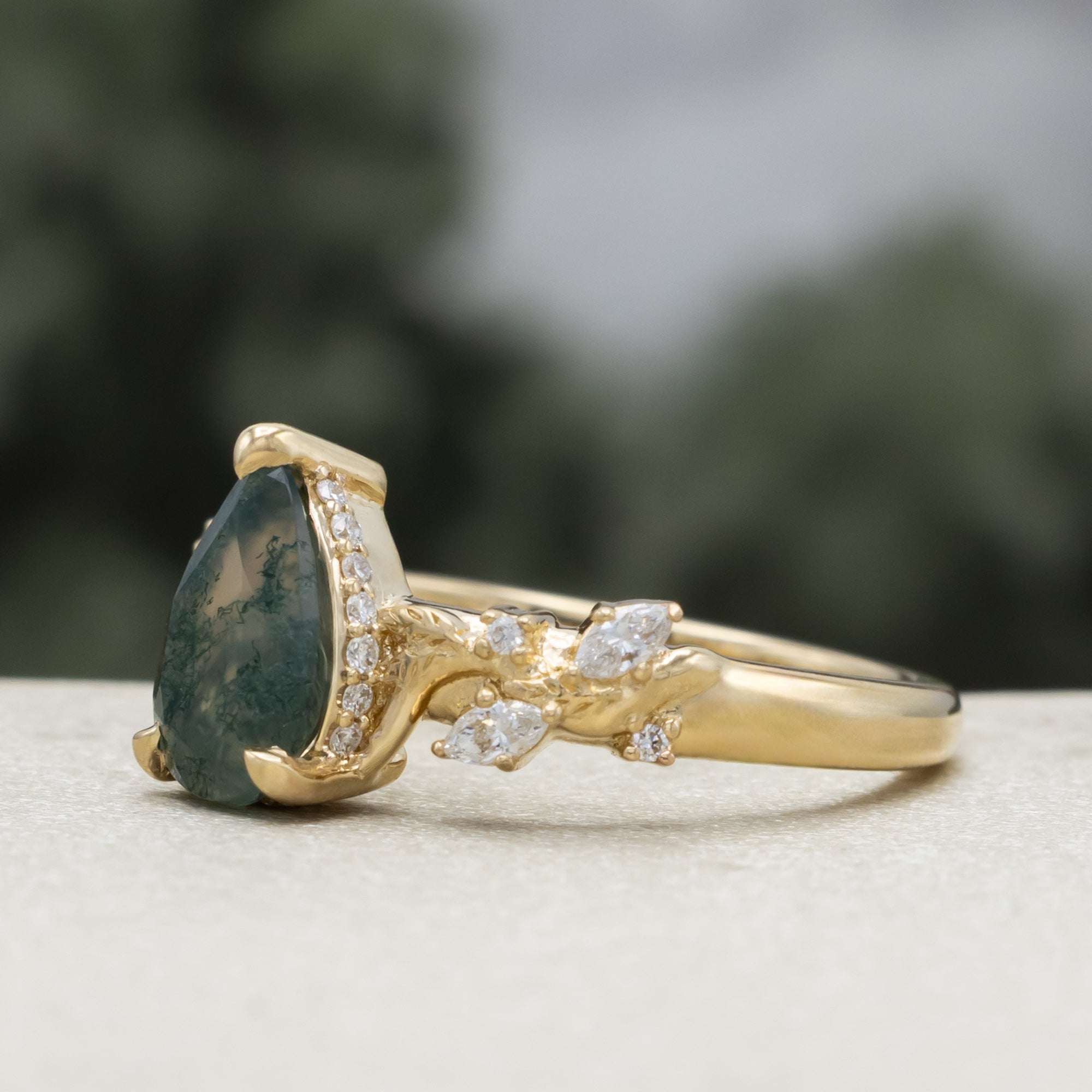 A gold ring featuring a pear cut moss agate flanked by delicate gold leaf-like branches with diamond accents, set against a blurred dark green background.