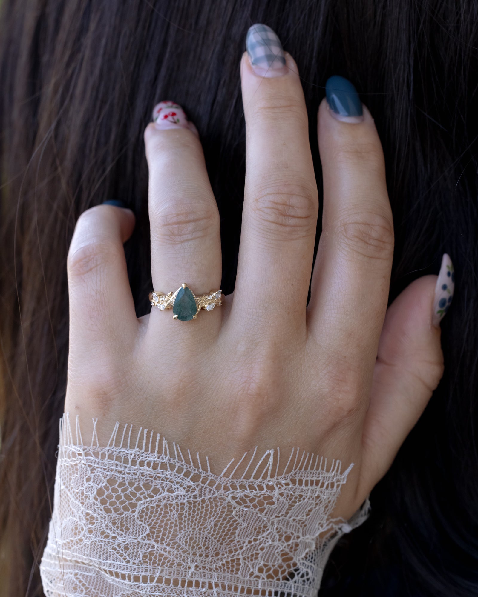 A close-up of a hand wearing a gold ring with a moss agate centerstone, resting against dark hair and a white lace cuff.