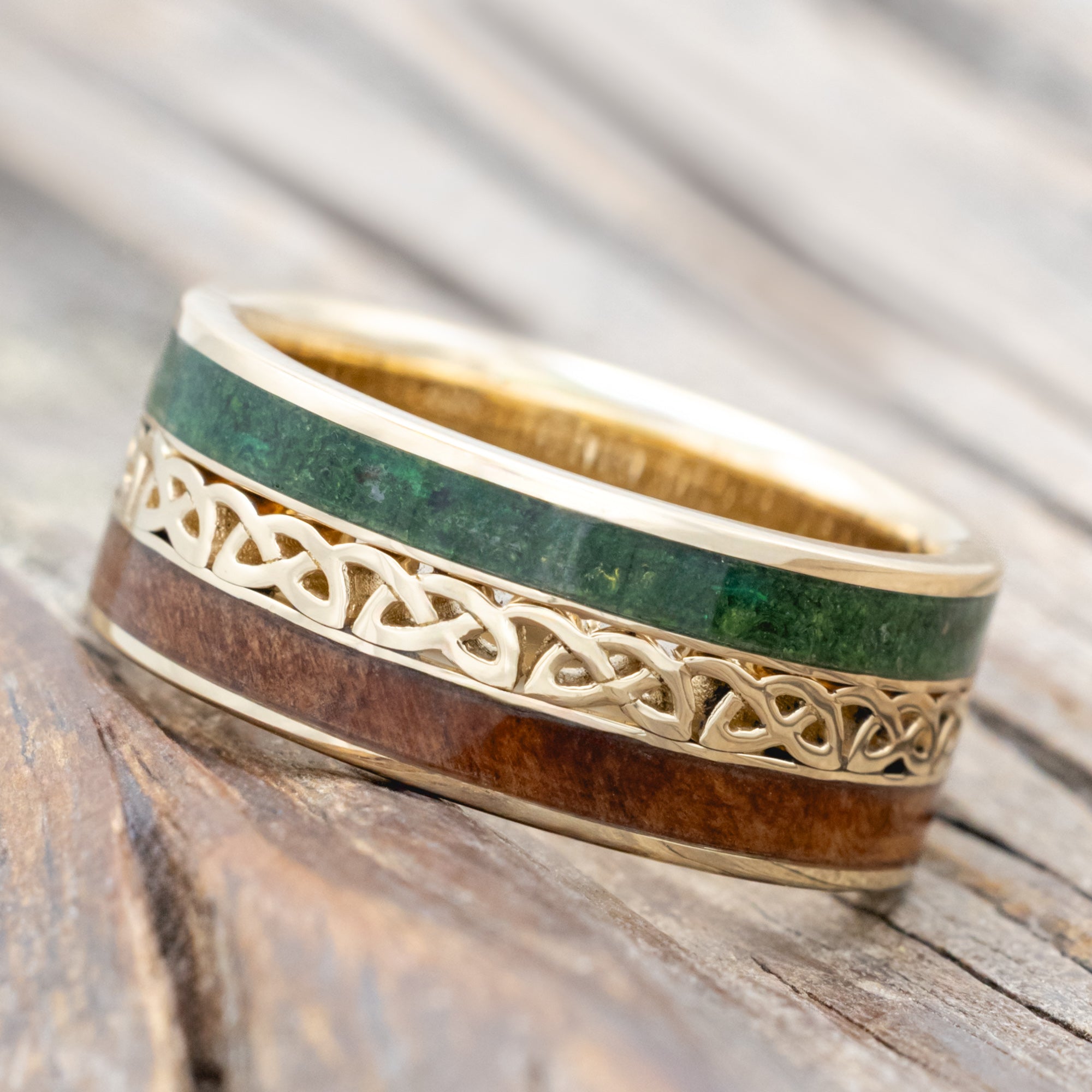 Gold ring with moss and redwood inlays with Celtic cutout on a wooden surface
