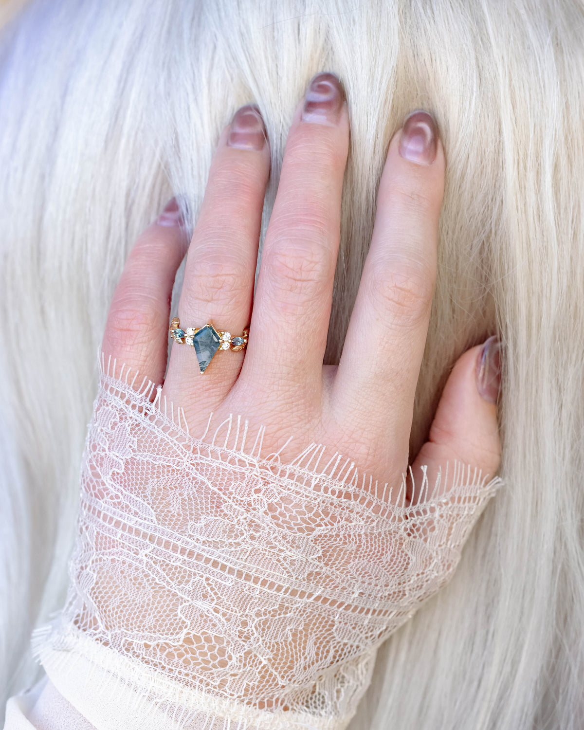 Hand wearing a ring with a moss agate center stone, in a lace sleeve, against a white background.