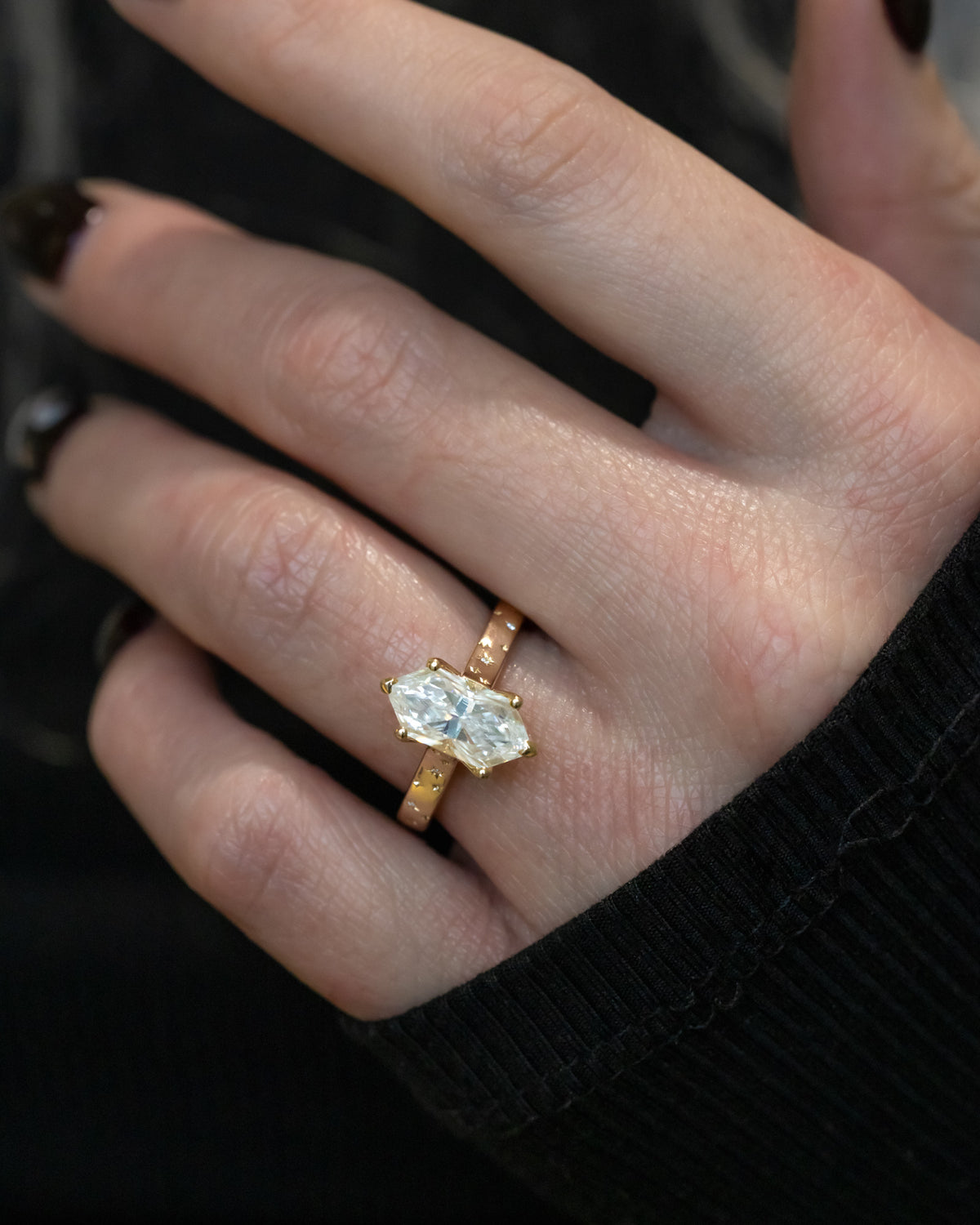 Close-up of a hand wearing a gold ring with a moissanite on a dark background