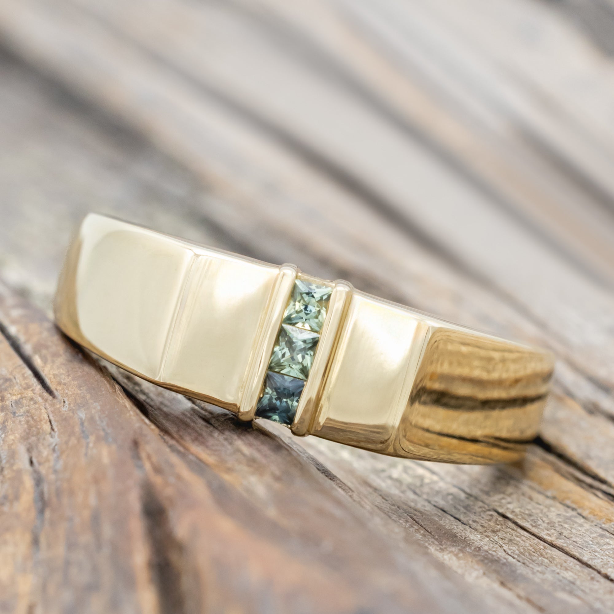 A polished yellow gold ring with a central channel set featuring light blue and green sapphires on a wooden surface.