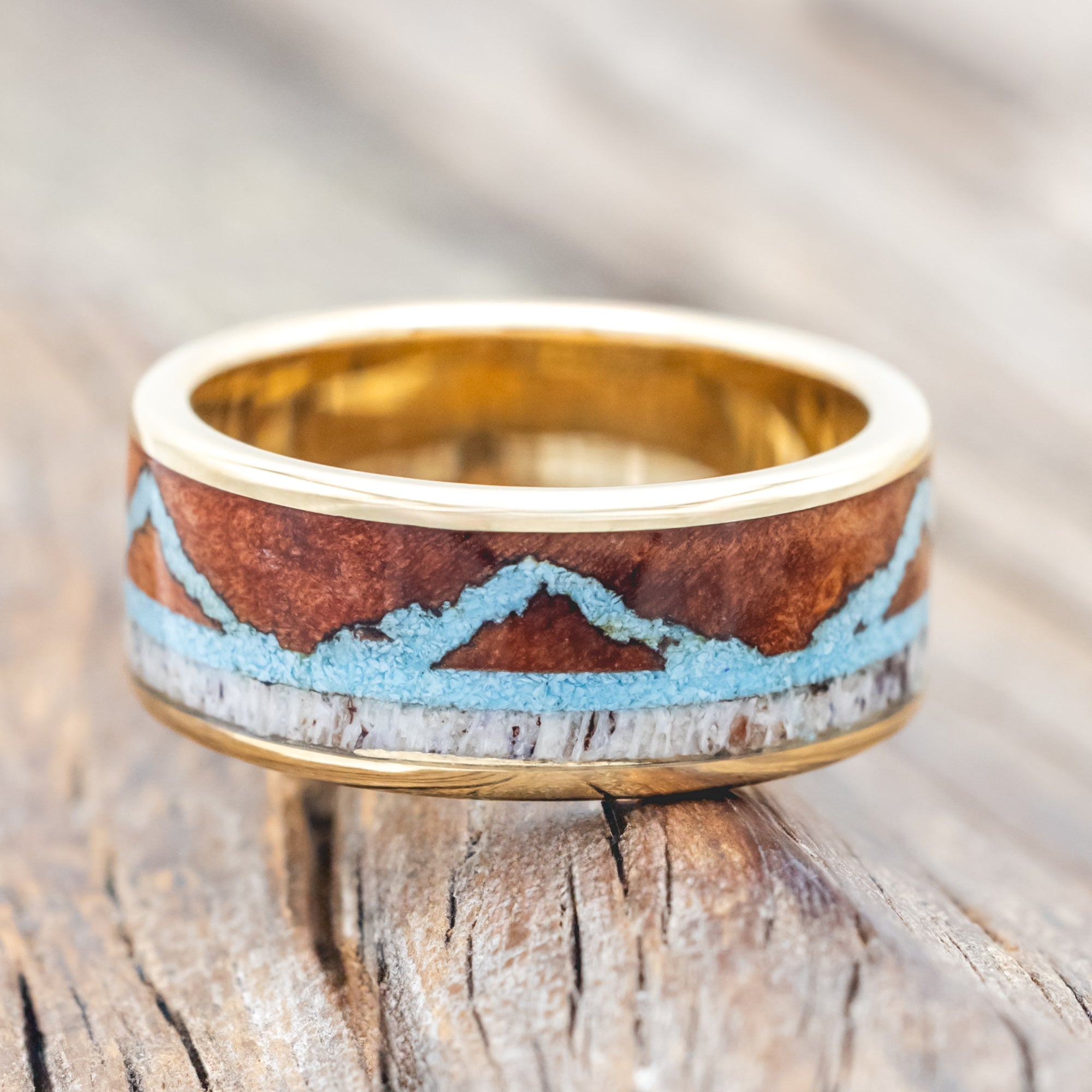 Yellow gold ring with whiskey barrel, antler, and turquoise mountains on a wooden surface