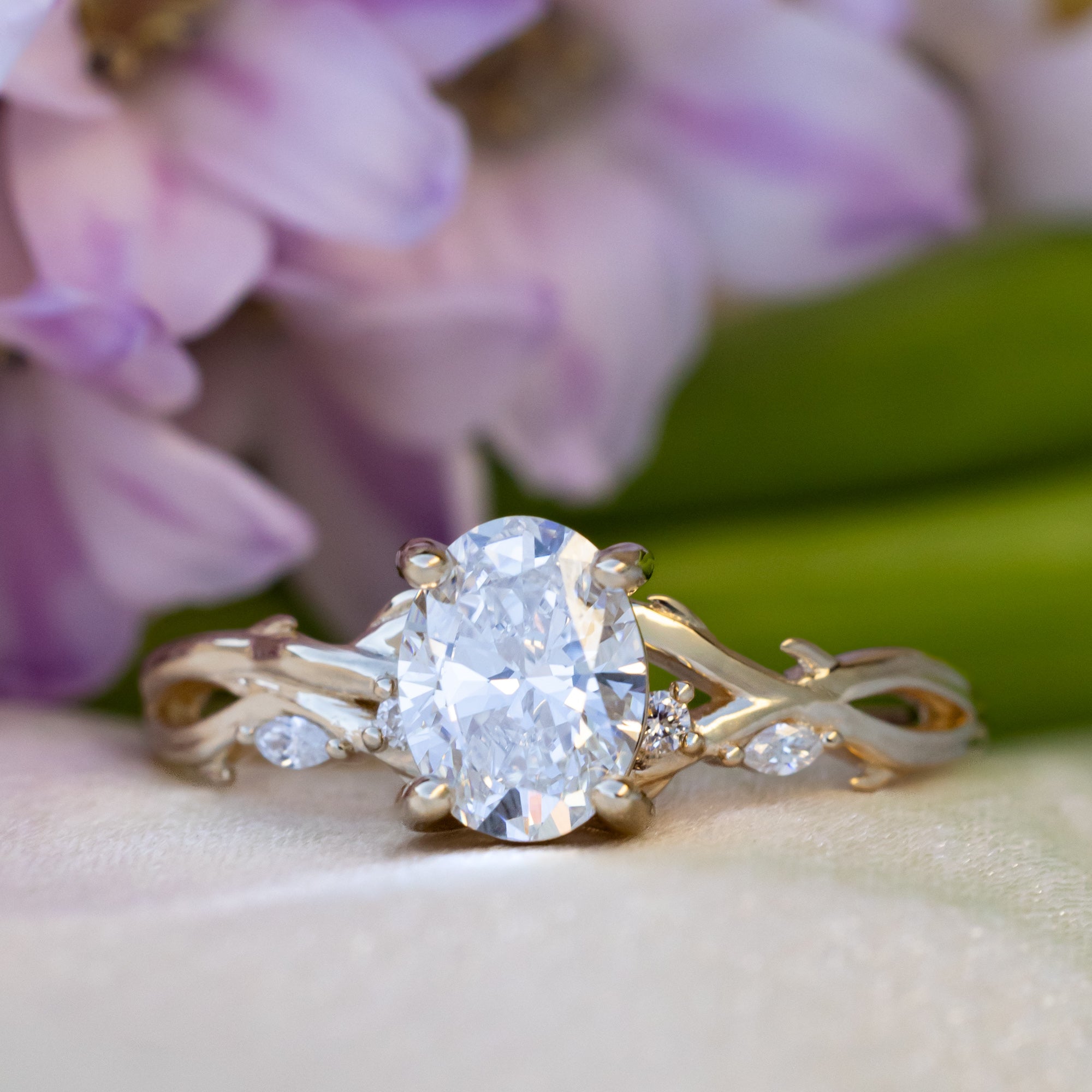 Gold ring with oval lab diamond center stone with diamond accents against a blurred floral background