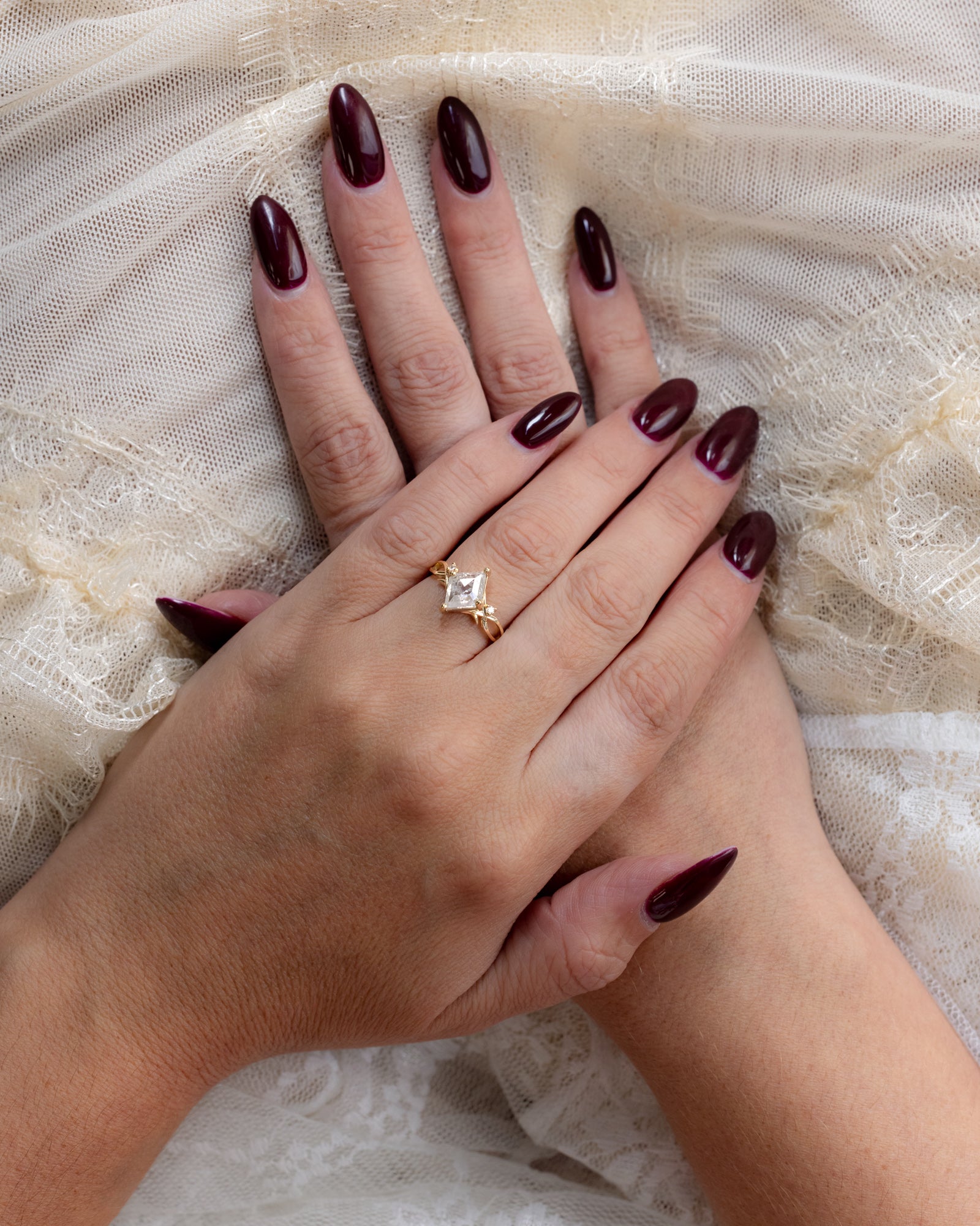 Hand with burgundy nail polish, wearing a gold salt & pepper diamond ring on a lace background