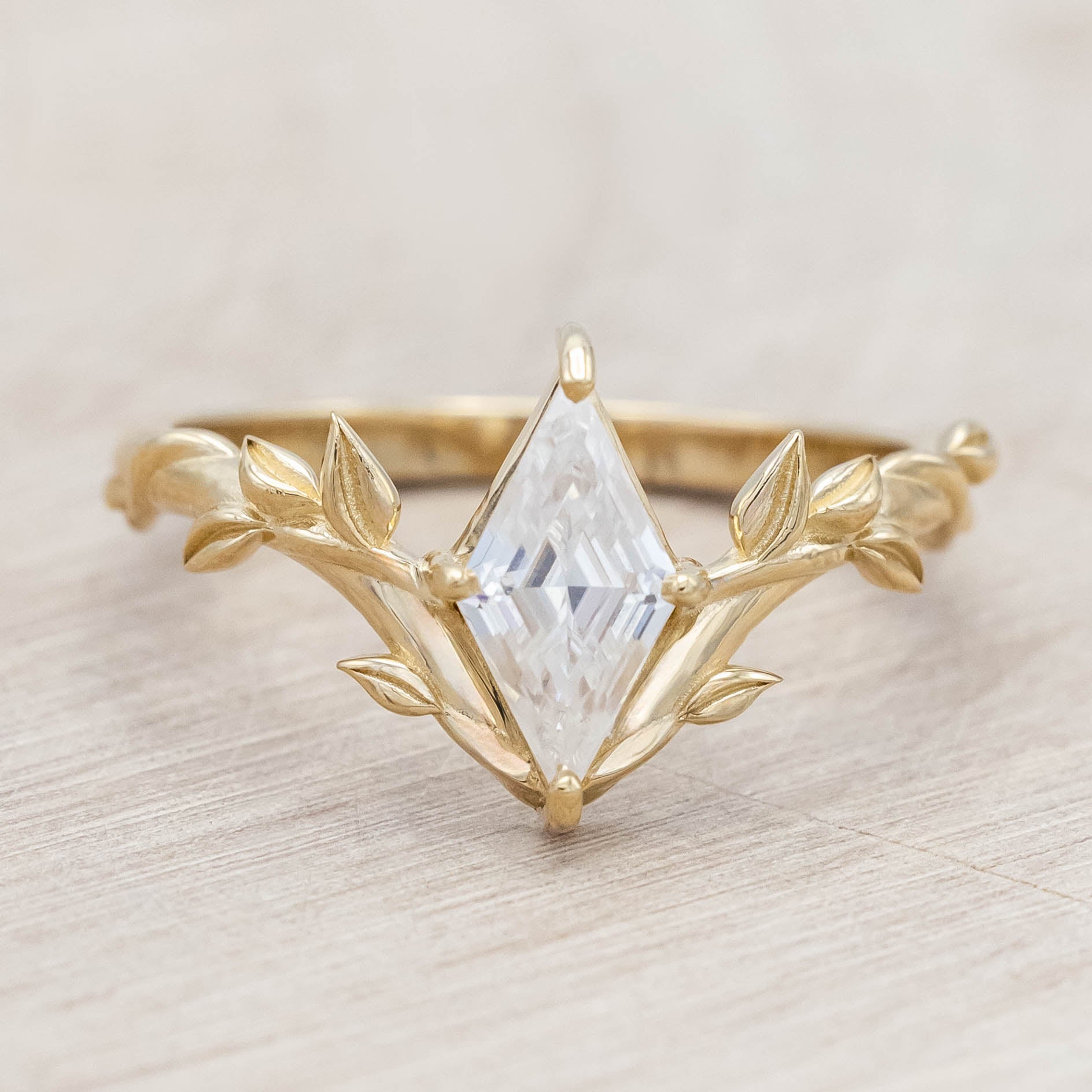 Gold vine-style ring with a lozenge Moissanite on a wooden stand against a neutral background