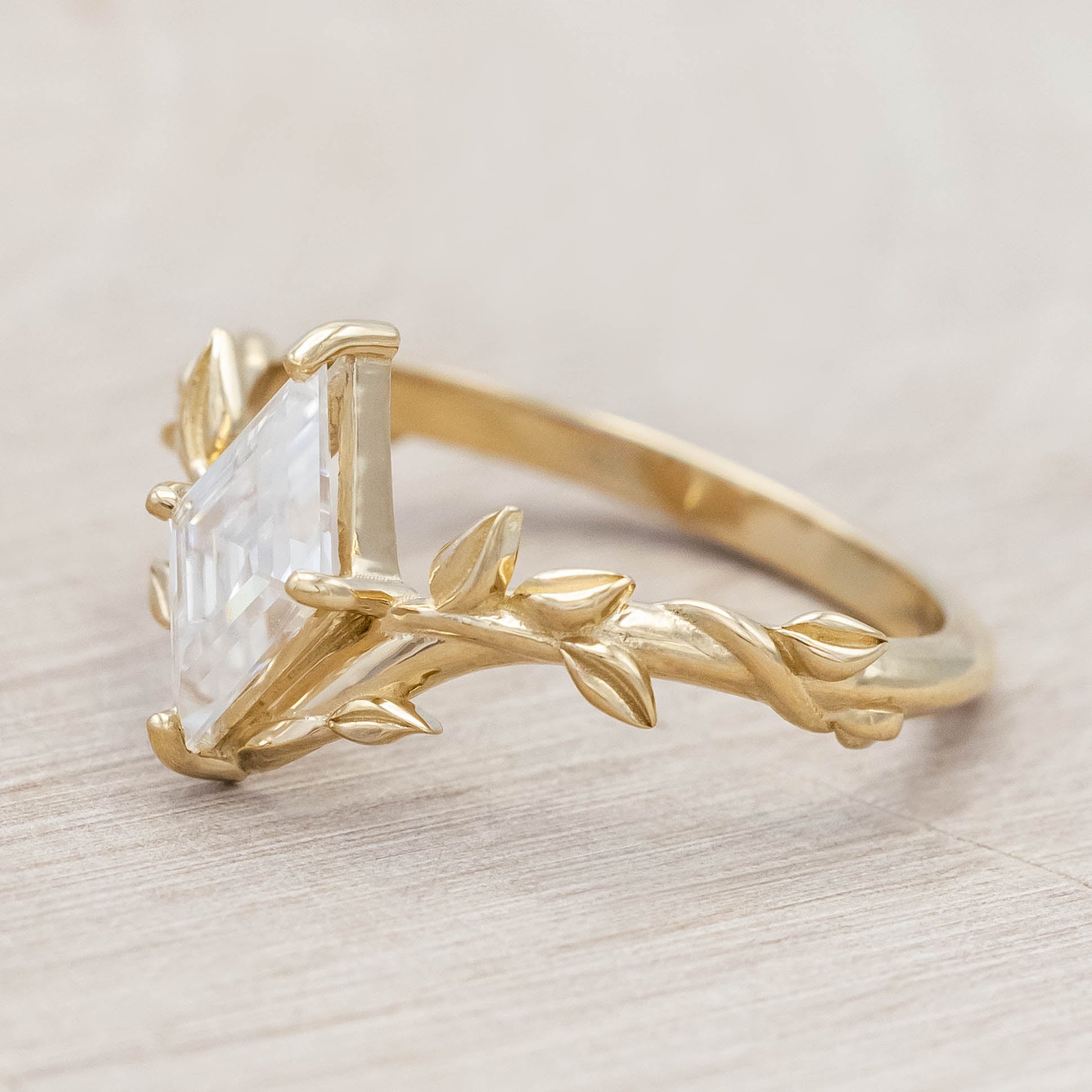 Gold vine-style ring with a lozenge Moissanite on a wooden stand against a neutral background