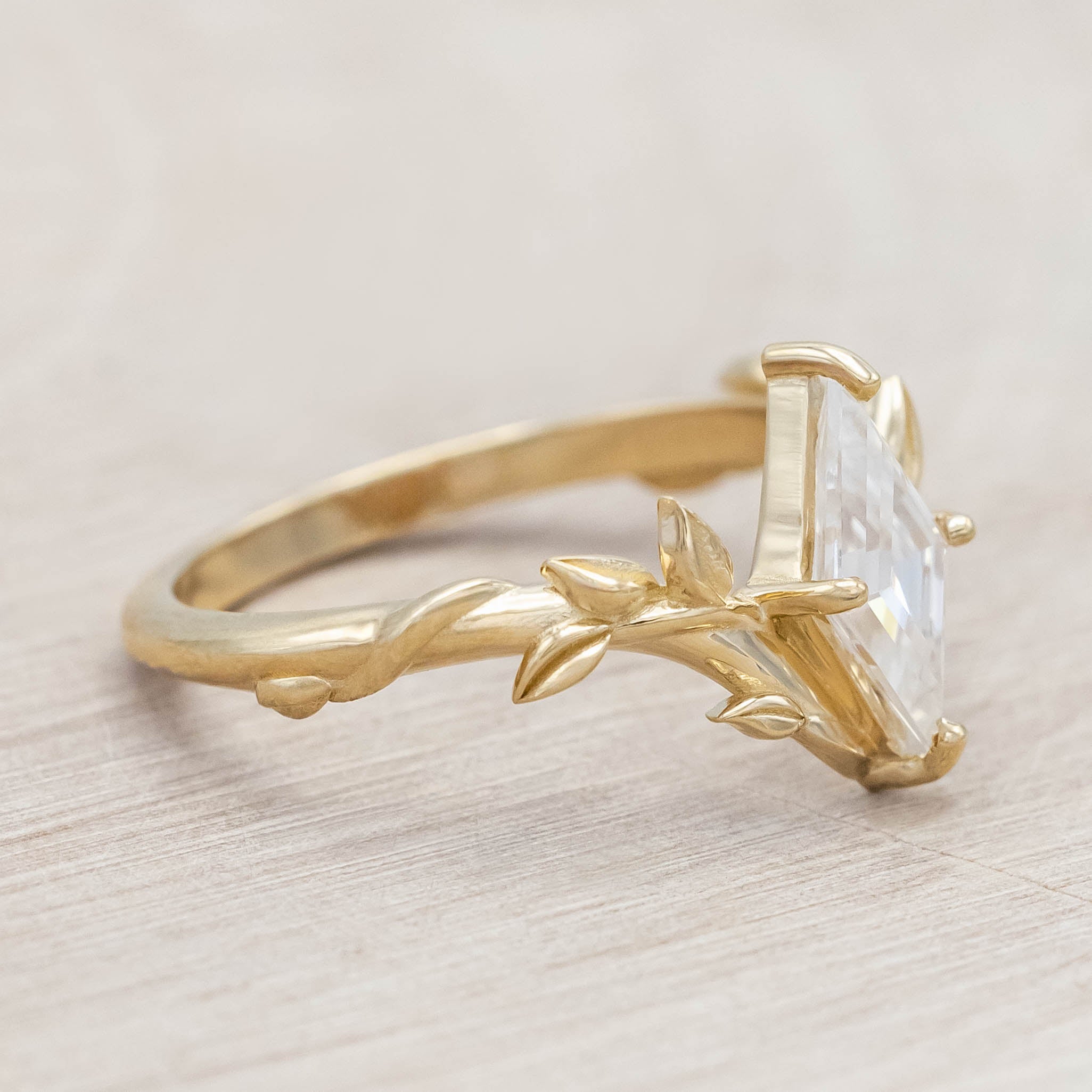 Gold vine-style ring with a lozenge Moissanite on a wooden stand against a neutral background
