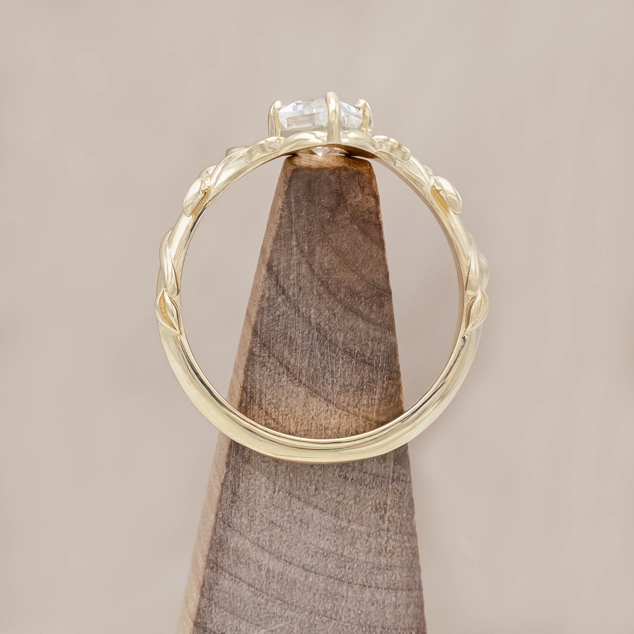 Gold vine-style ring with a lozenge Moissanite on a wooden stand against a neutral background