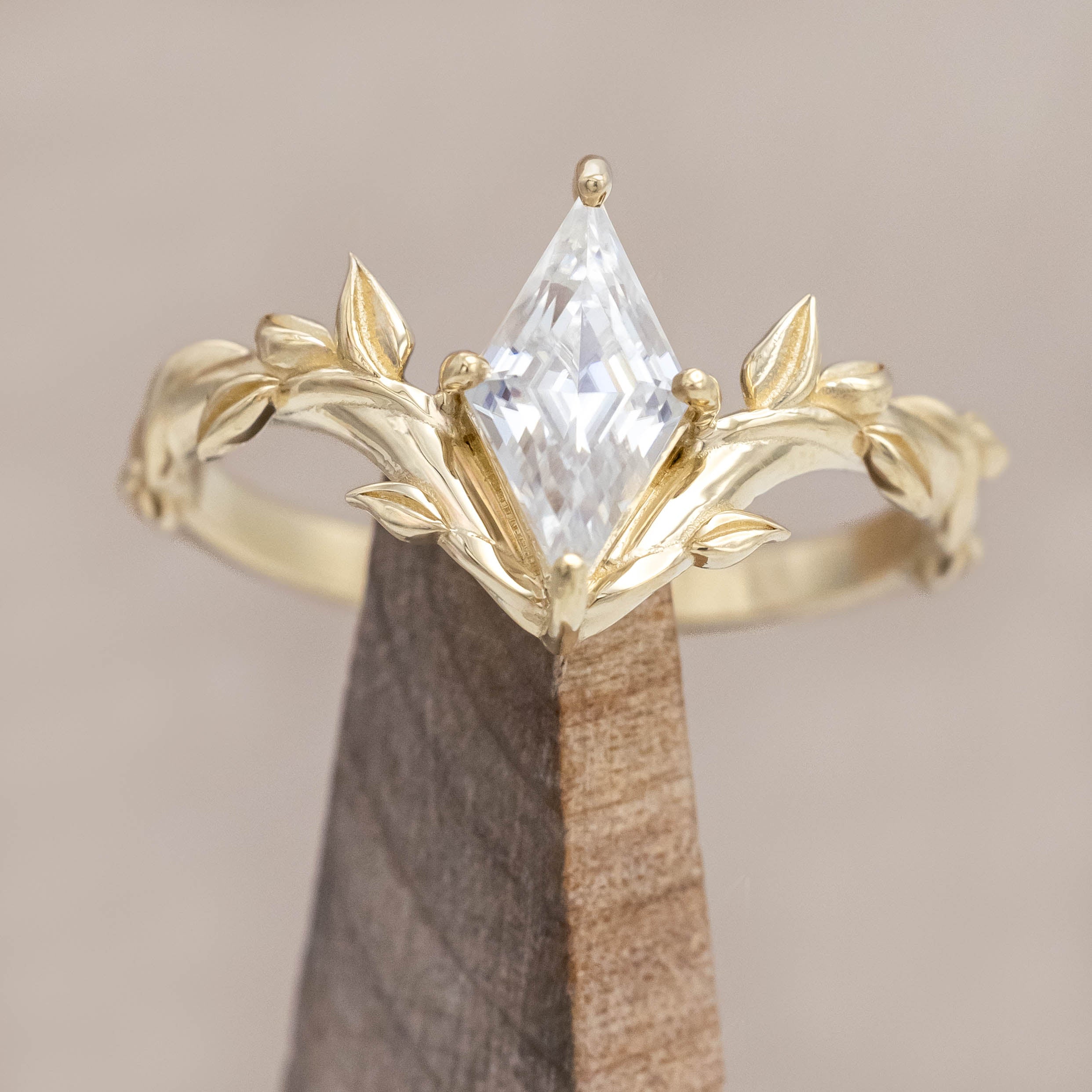 Gold vine-style ring with a lozenge Moissanite on a wooden stand against a neutral background