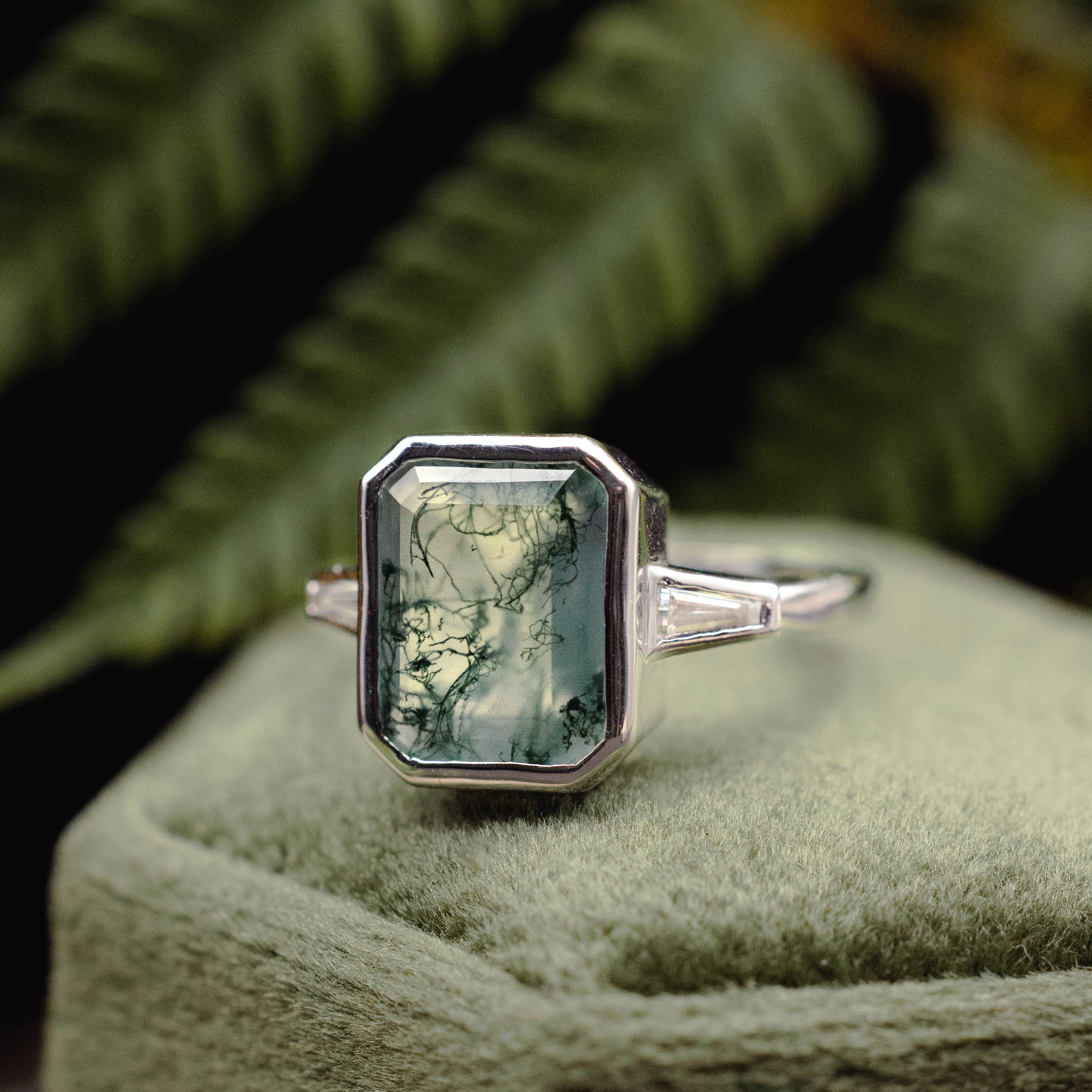 White gold ring with moss agate on a green cushion with ferns in the background
