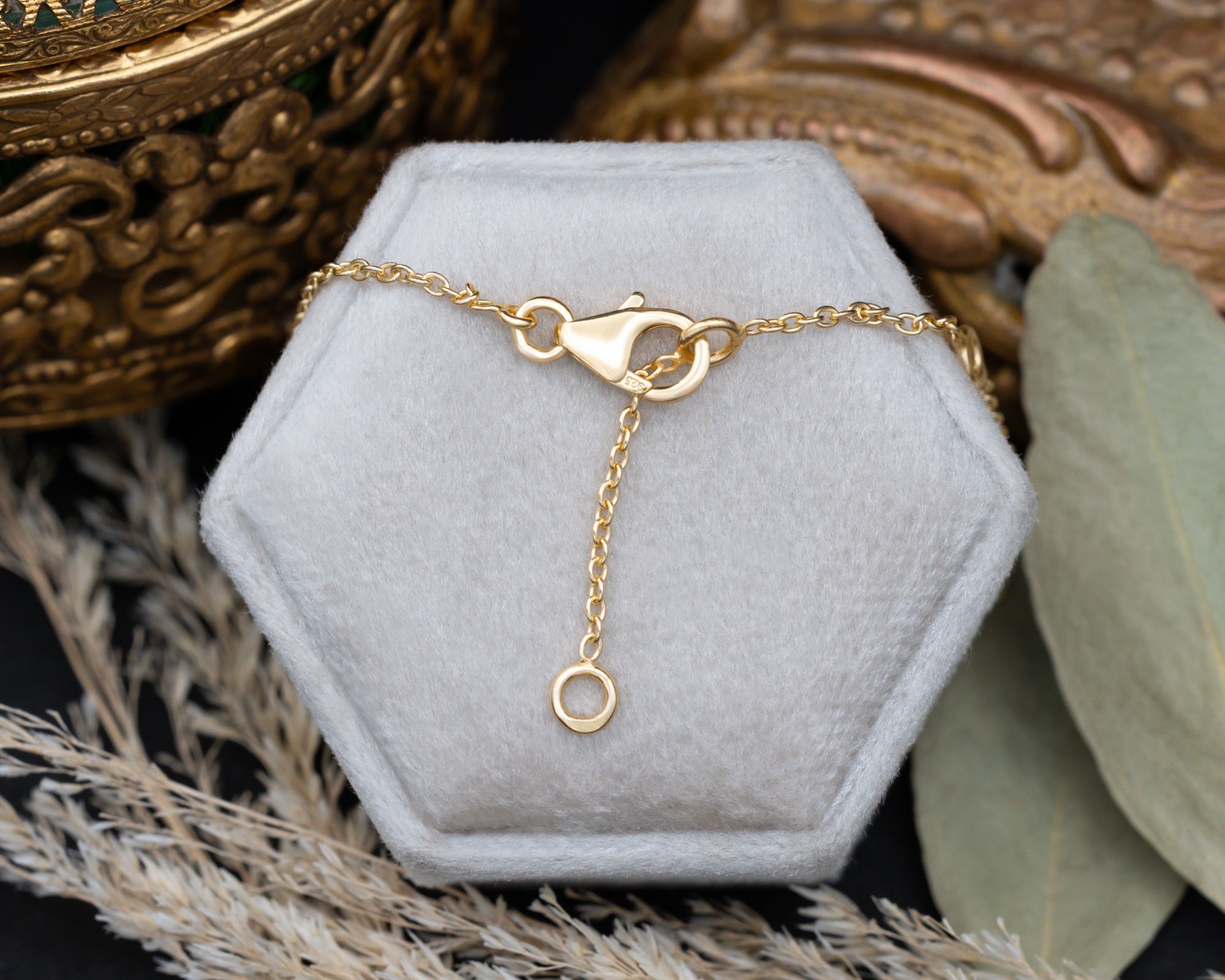 A delicate gold chain necklace with a lobster clasp, draped over two hexagonal white jewelry display boxes on a dark surface, styled with dried leaves and a decorative gold bowl.