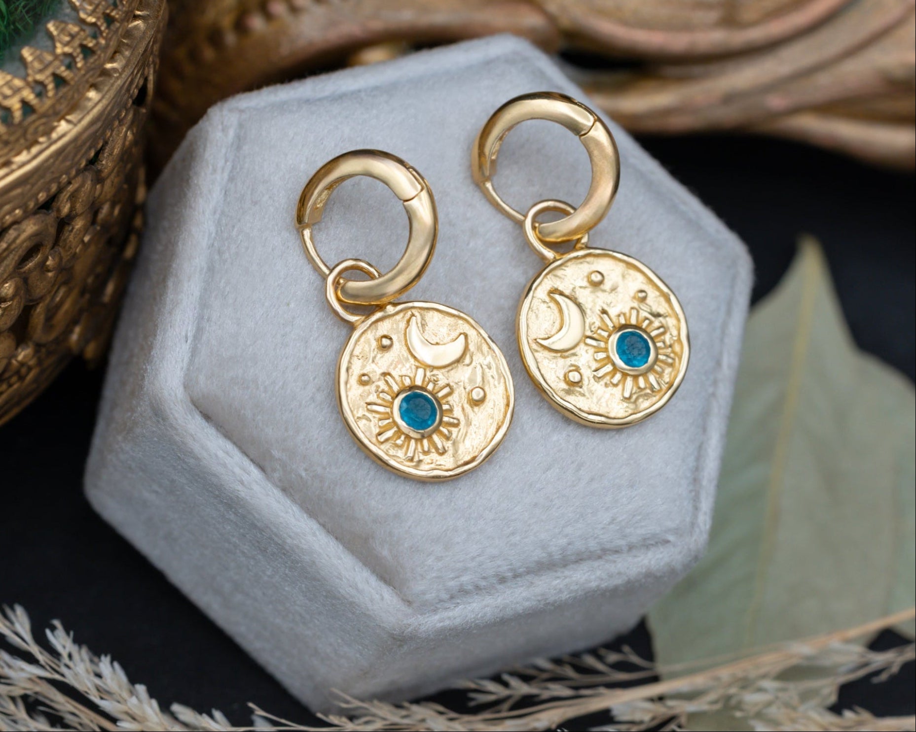 A pair of gold crescent moon and sun earrings with blue apatite gemstone accents , displayed on a velvet jewelry box surrounded by decorative dried grass