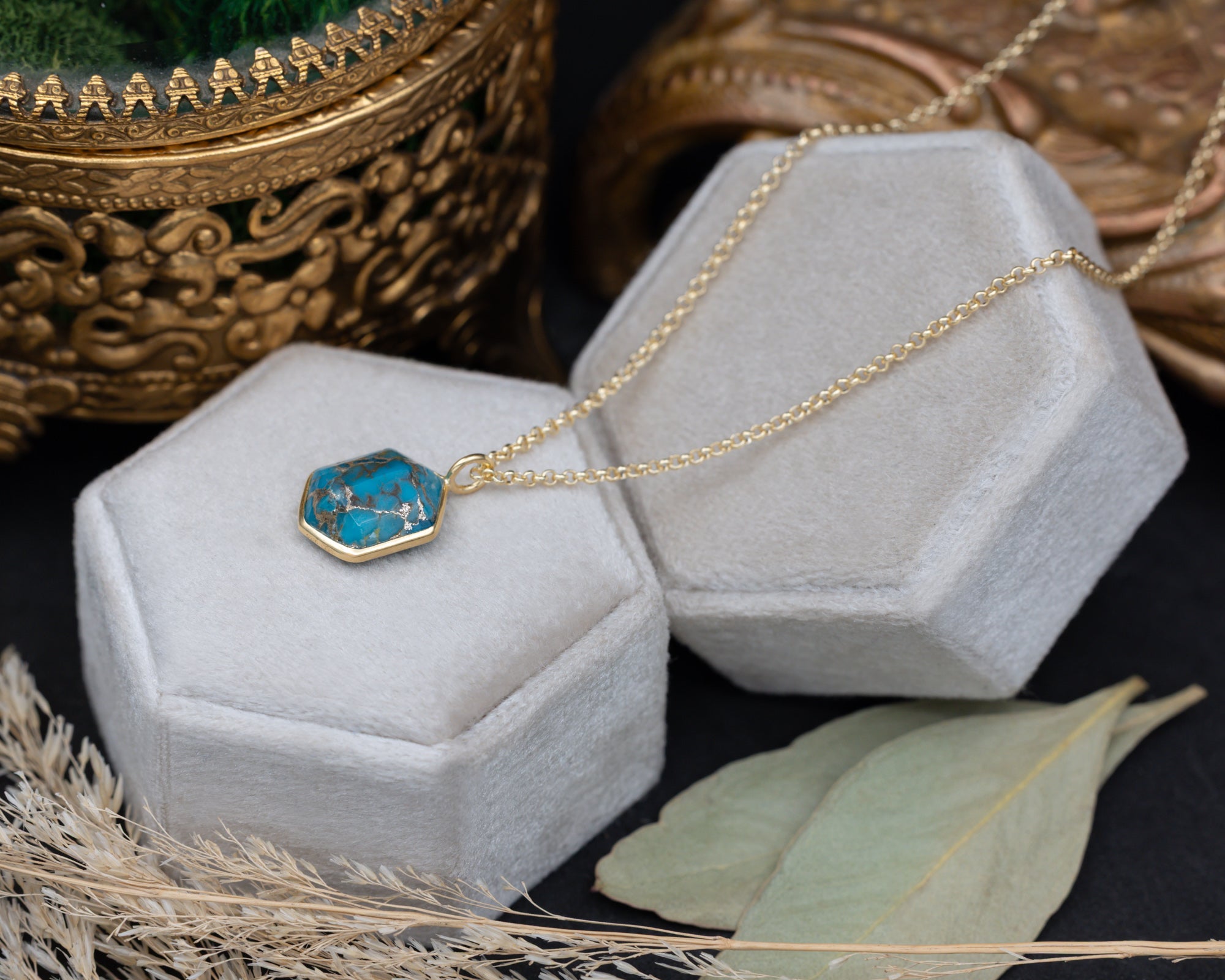 A turquoise pendant on a gold chain against a white cushion with dried flowers in the background