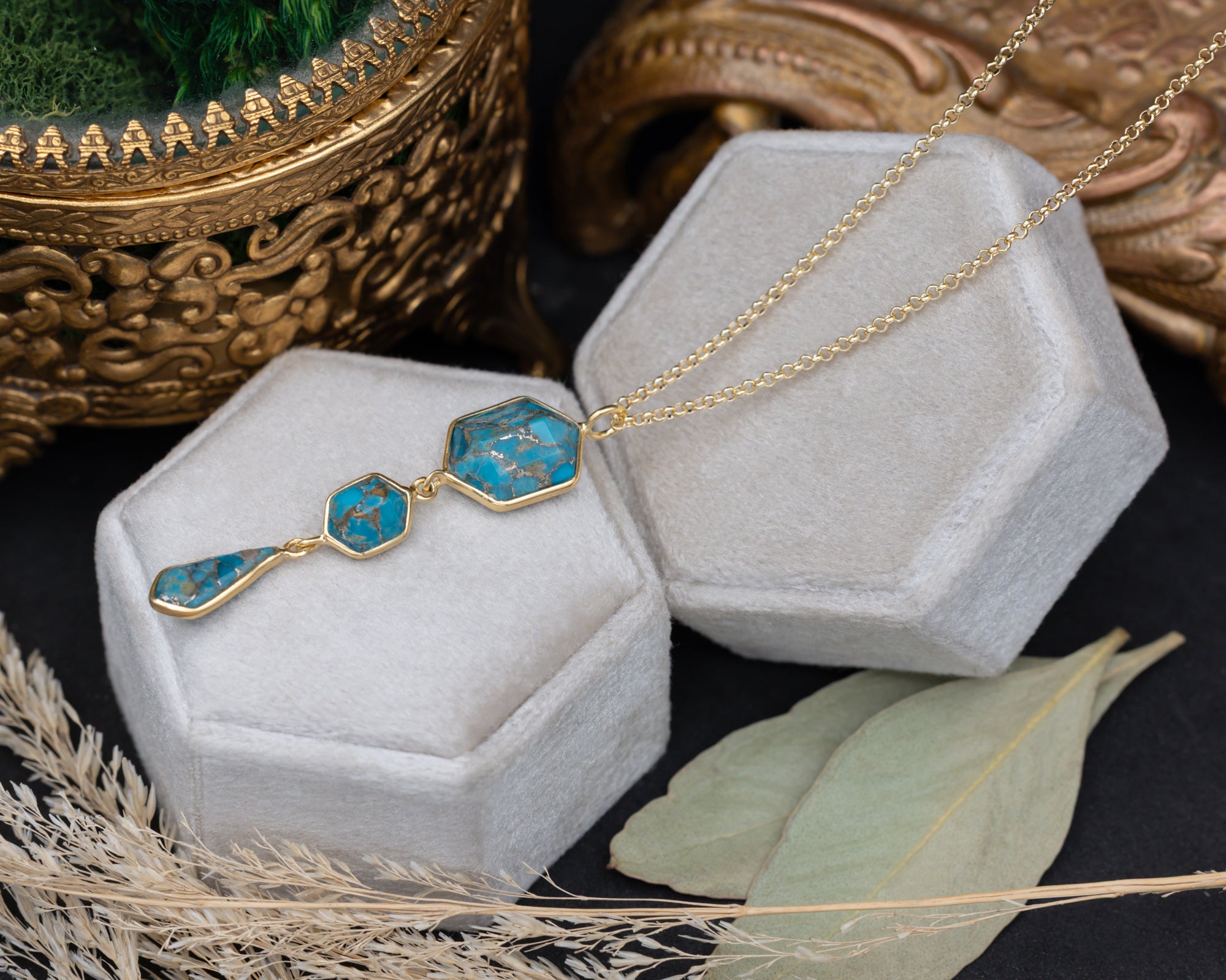 A gold pendant necklace with three turquoise stones on a gold chain, sitting on a white cushion with dried flowers in the background