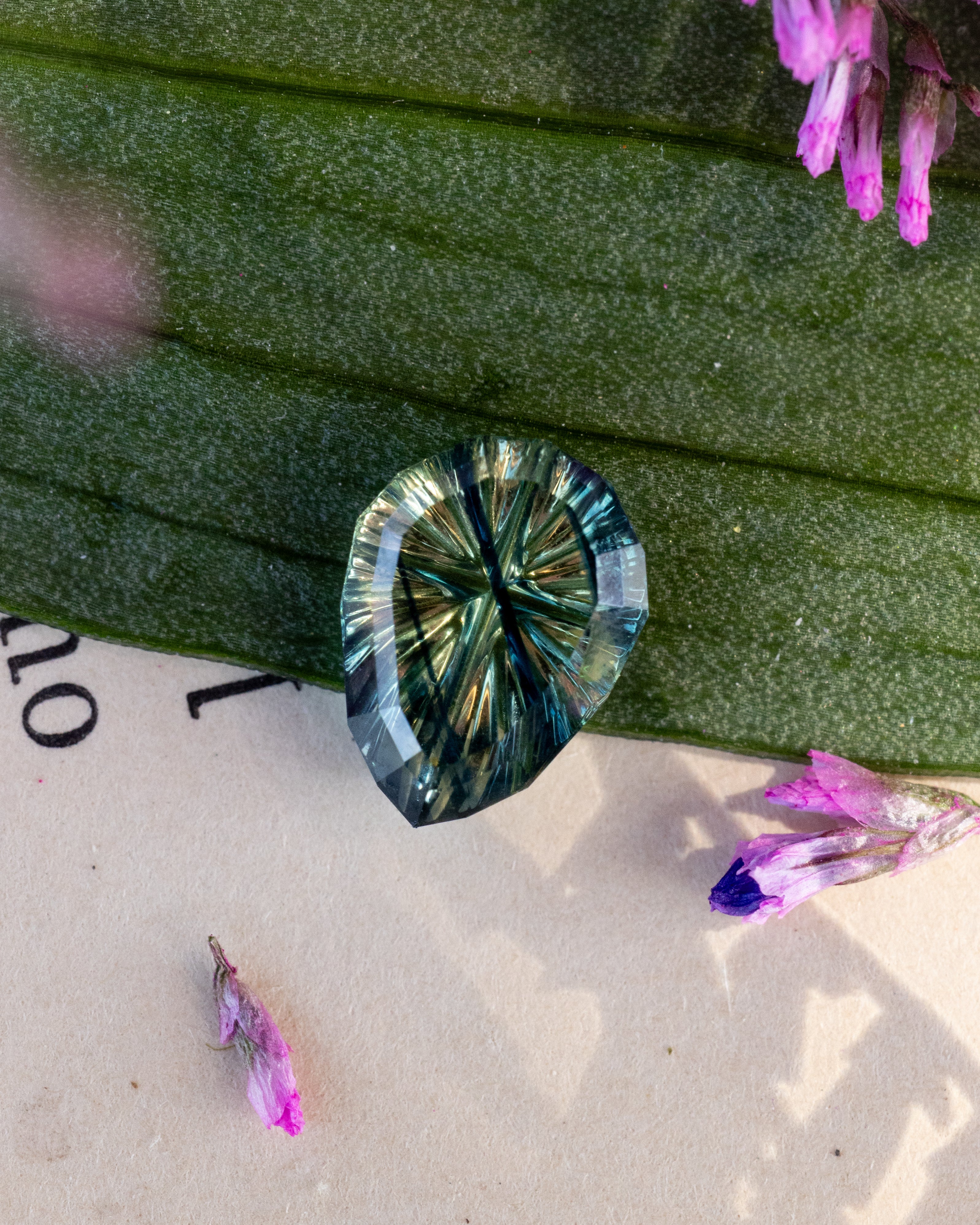 A faceted pear sapphire with a starburst pattern of green, blue, and gold iridescence, resting on a textured green leaf with small pink flowers on a beige surface.