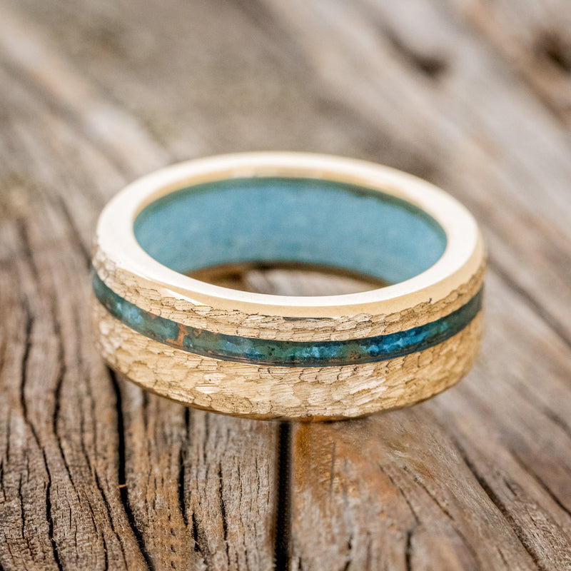 Shown here is "Vertigo", a custom, handcrafted men's wedding ring featuring an offset copper patina inlay and a turquoise lining on a hammered 14K gold band, laying flat. Additional inlay options are available upon request.