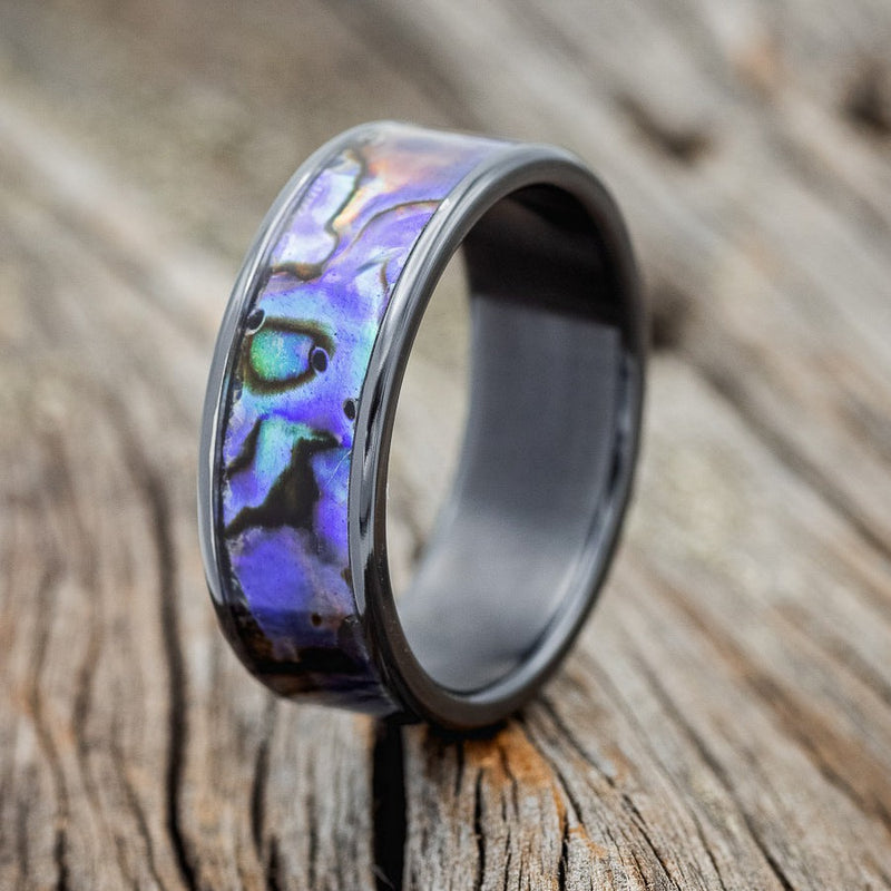 Shown here is "Rainier", a handcrafted men's wedding ring featuring paua shell inlay and shown here on a fire-treated black zirconium band, upright facing left. Additional inlay options are available upon request.