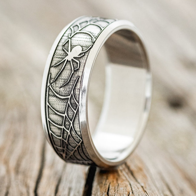 Shown here is "Legacy", a handcrafted, embossed men's wedding ring featuring a spider and spider web engraving in a channel-style band, upright facing left. It can be customized to feature just about any embossed design you can dream up.