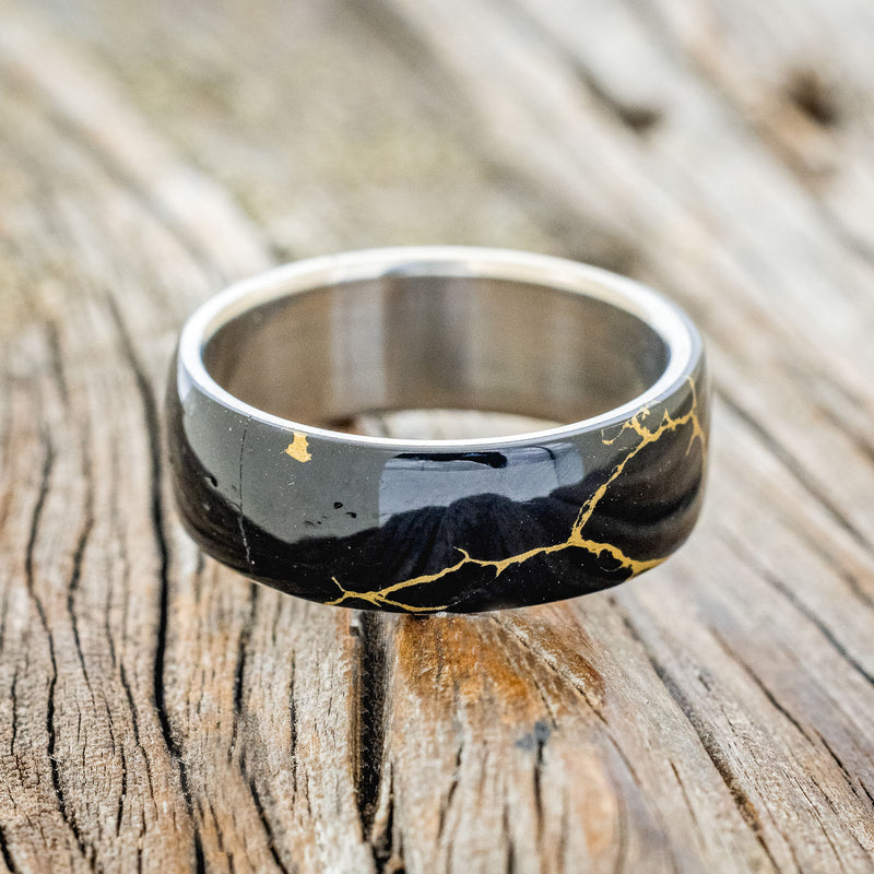 Shown here is "Haven", a custom, handcrafted men's wedding ring featuring a black and gold TruStone overlay on a titanium band, laying flat. Additional overlay options are available upon request.