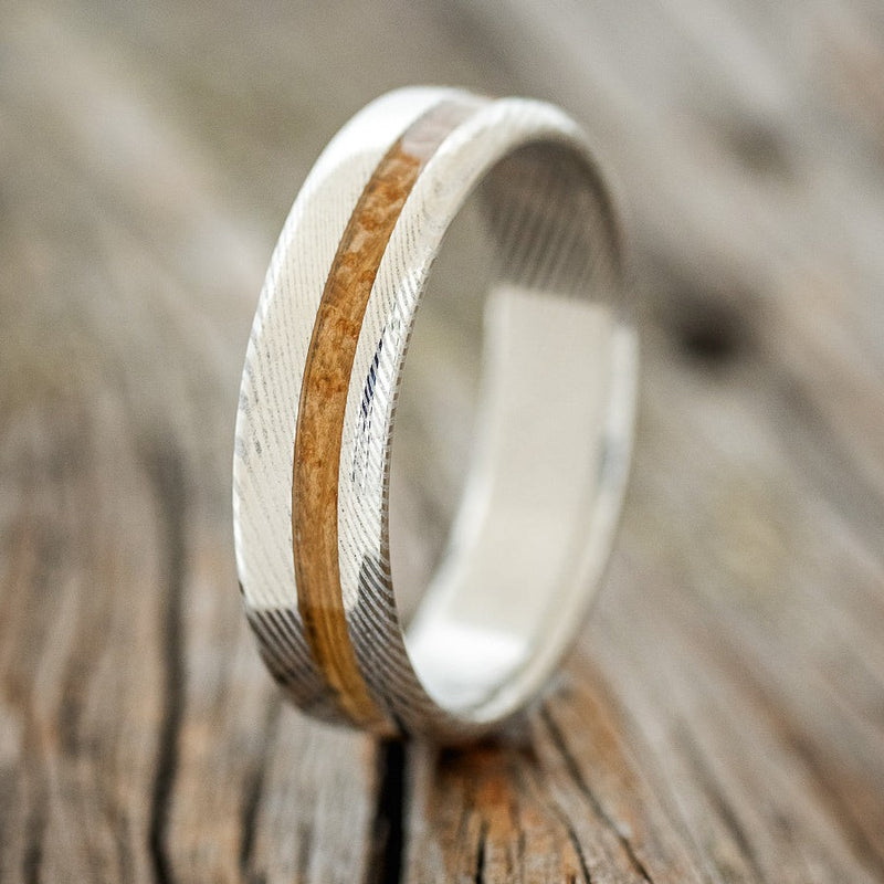 Shown here is "Vertigo", a custom, handcrafted men's wedding ring featuring a whiskey barrel inlay, shown here set on a Damascus steel band, upright facing left. Additional inlay options are available upon request.