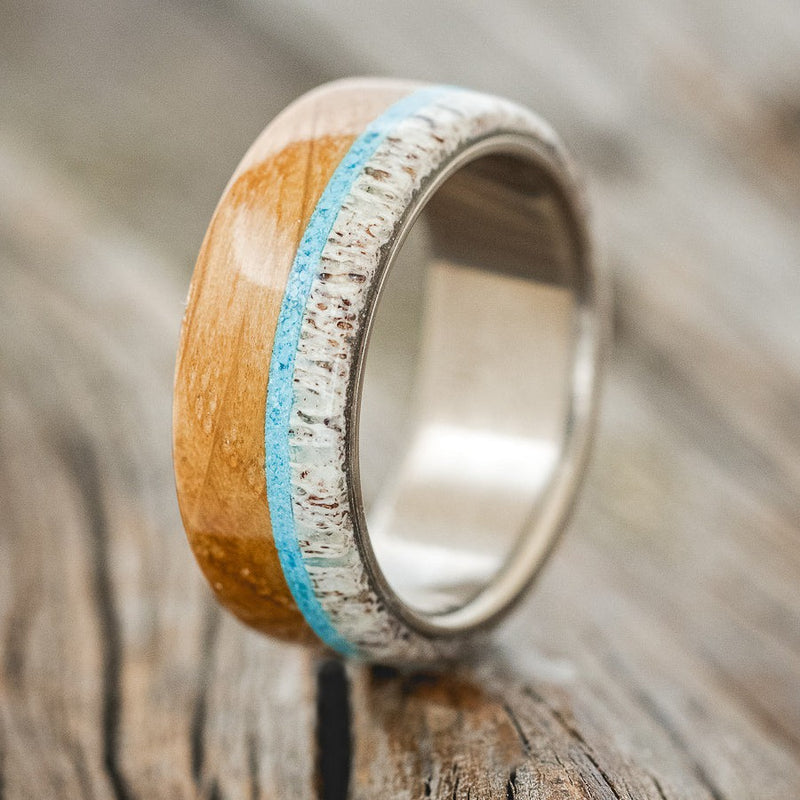 Shown here is "Banner", a custom, handcrafted men's wedding ring featuring whiskey barrel oak and antler overlay with a turquoise inlay on a titanium band, upright facing left. Additional inlay options are available upon request.