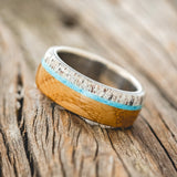Shown here is "Banner", a custom, handcrafted men's wedding ring featuring whiskey barrel oak and antler overlay with a turquoise inlay on a titanium band, tilted left. Additional inlay options are available upon request.