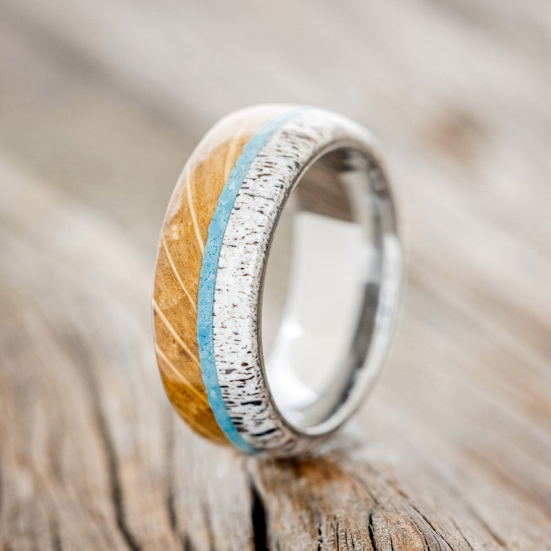 Shown here is "Banner", a custom, handcrafted men's wedding ring featuring whiskey barrel oak and antler overlay with a turquoise inlay, upright facing left. Additional inlay options are available upon request.