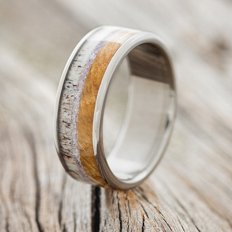 Shown here is "Rainier", a custom, handcrafted men's wedding ring featuring an authentic whiskey barrel wood, charoite, and elk antler inlays, shown here on a titanium band, upright facing left. Additional inlay options are available upon request.