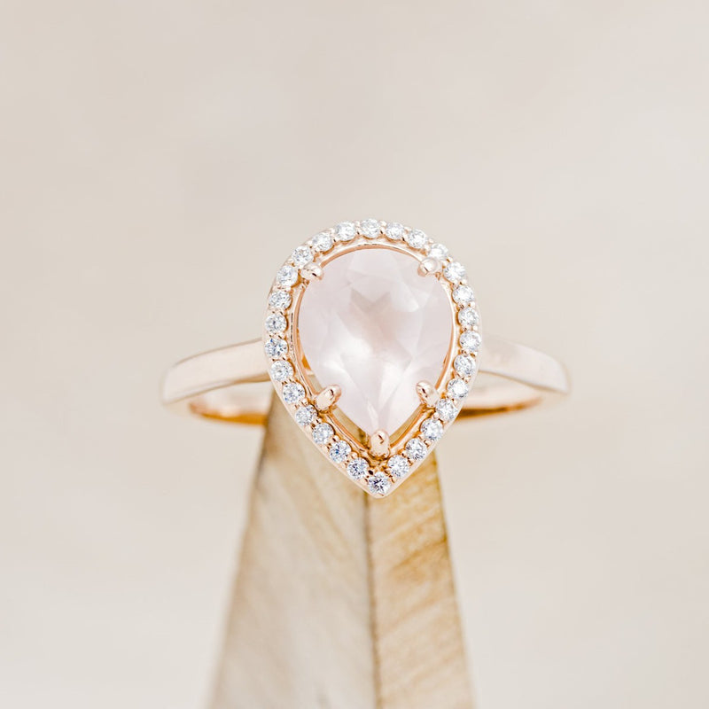 Shown here is "Clariss", a rose quartz women's engagement ring with a diamond halo, on stand front facing. Many other center stone options are available upon request. 