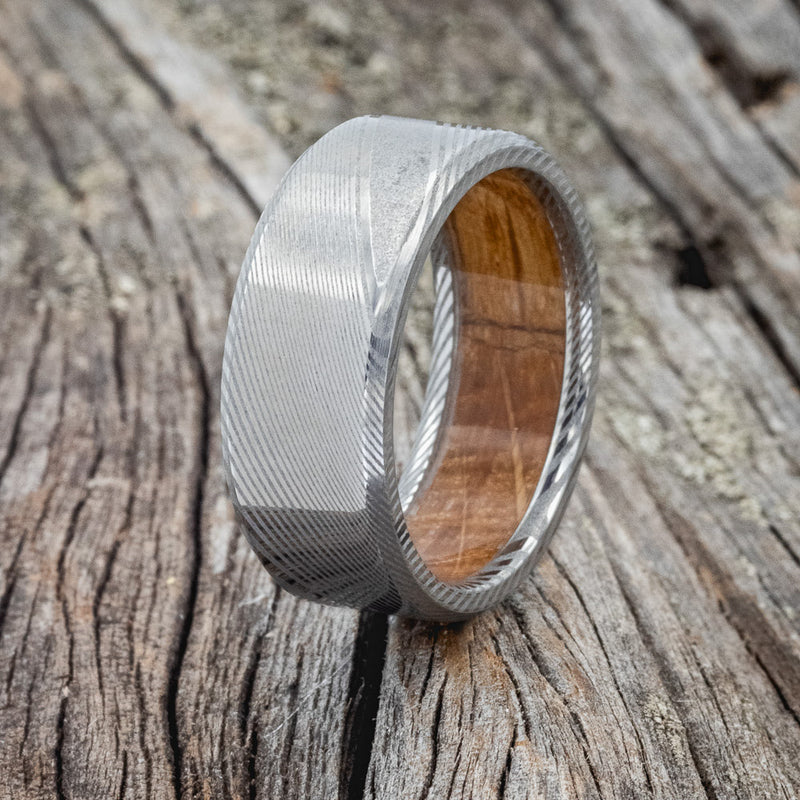 Shown here is a custom, handcrafted men's wedding ring featuring a hand-turned Damascus Steel wedding band lined with oak from a whiskey barrel, upright facing left. Additional inlay options are available upon request.