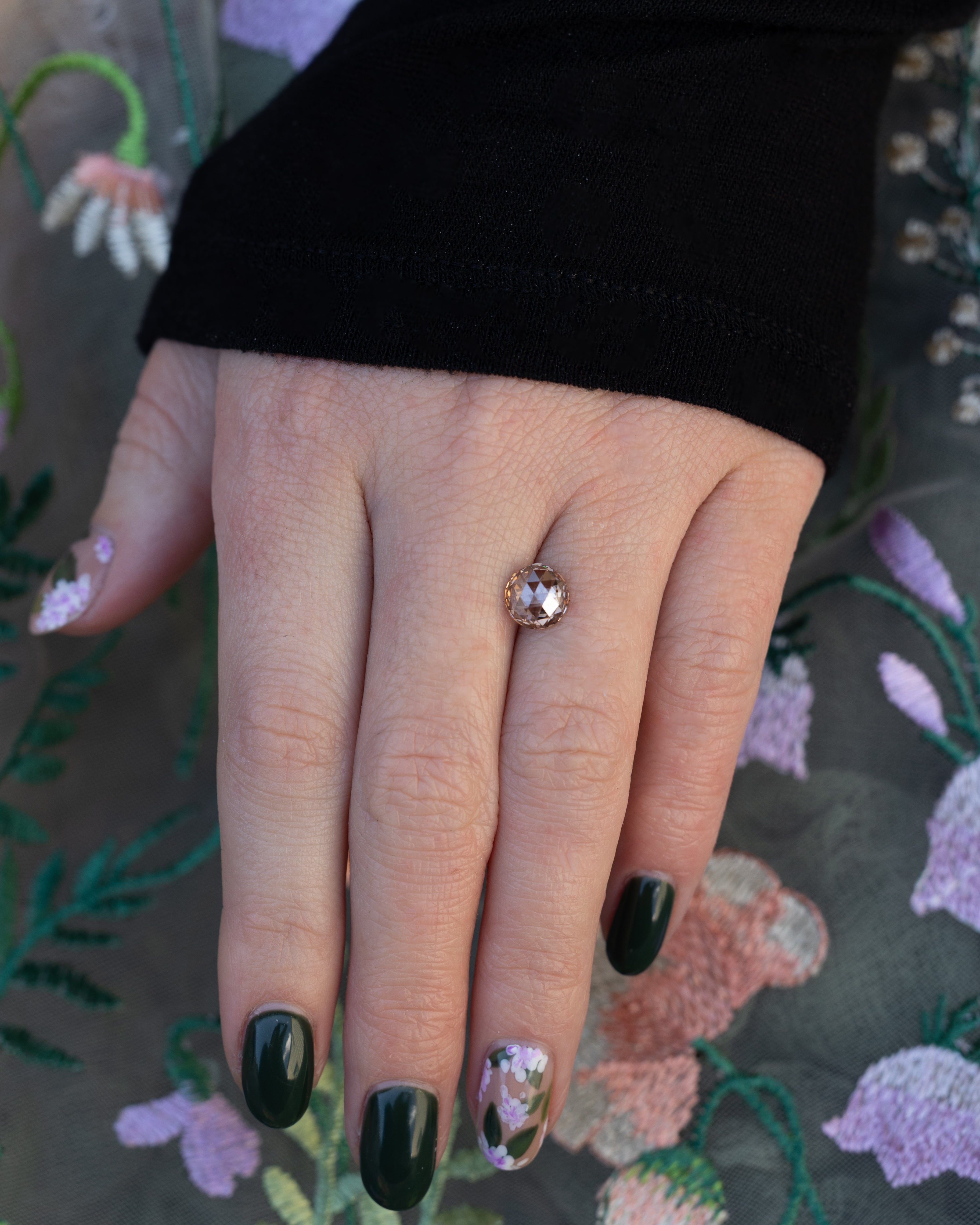 Hand holding a round champagne diamond with a black sleeve against a floral background.