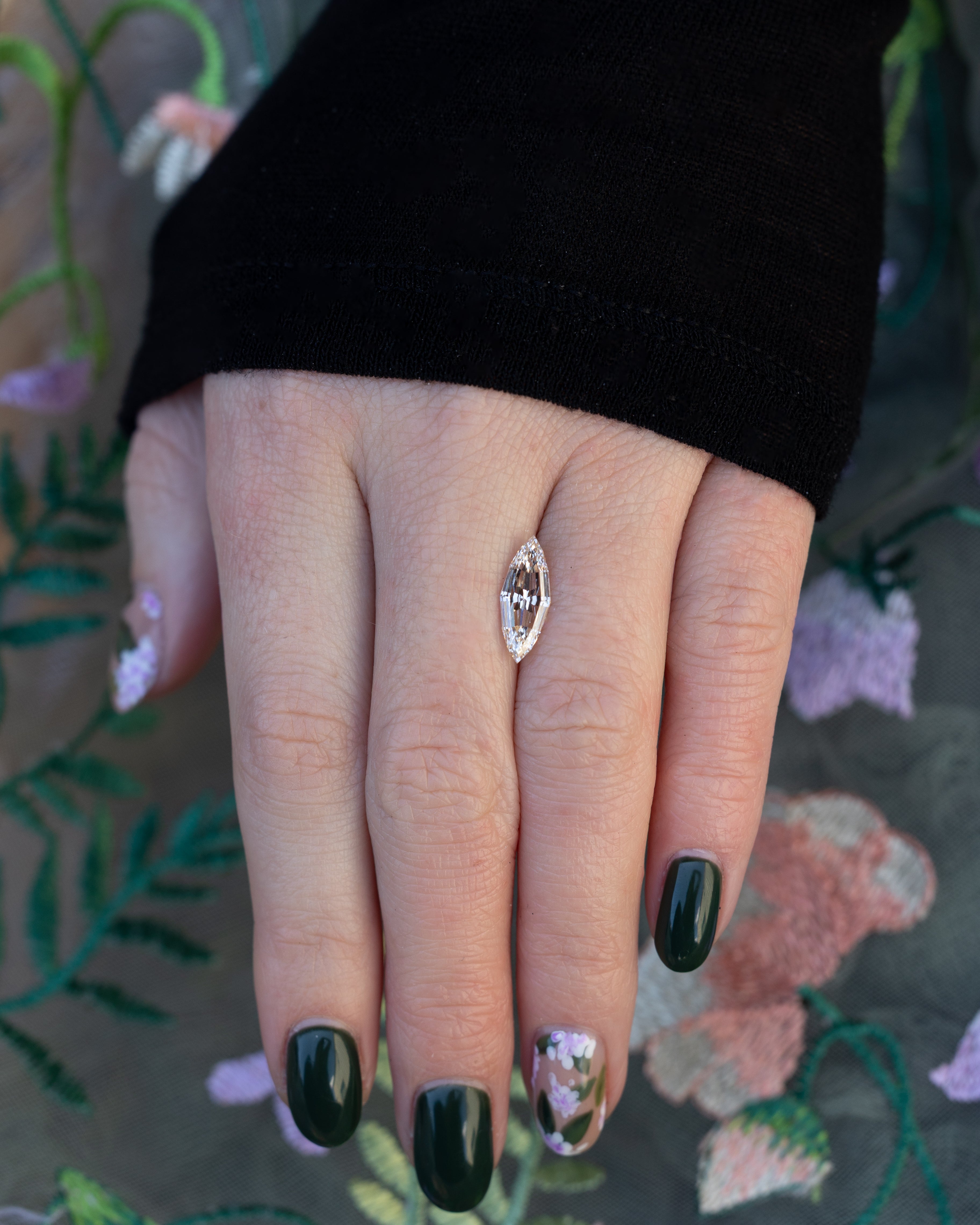 Hand holding a marquise champagne diamond with a black sleeve against a floral background.