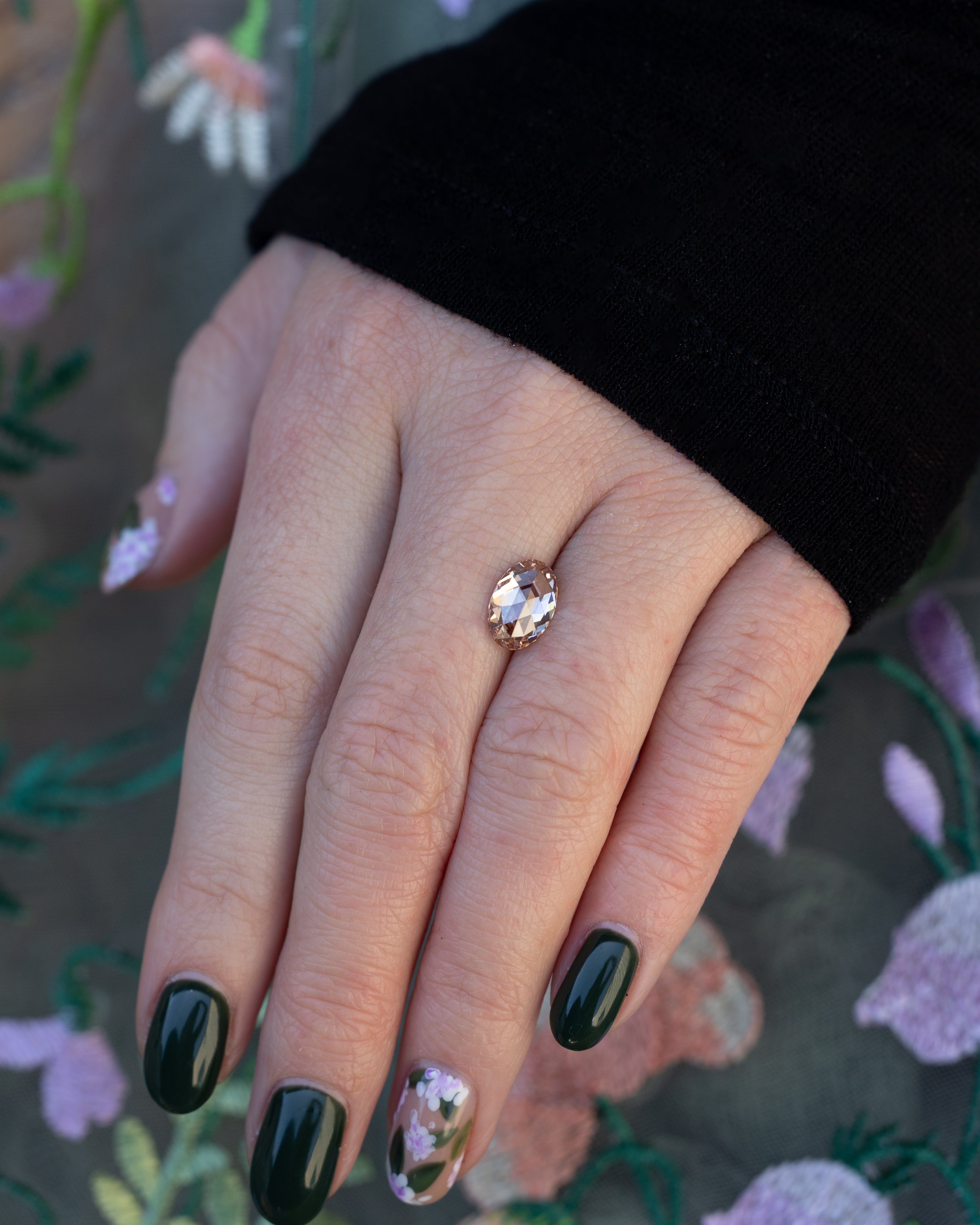 Hand holding an oval champagne diamond with a black sleeve against a floral background.