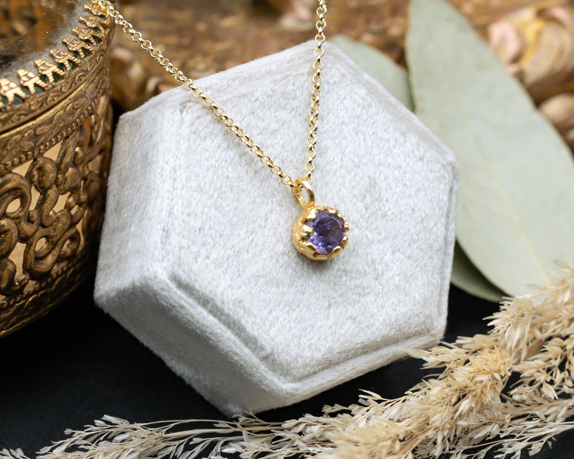 Gold necklace with a purple amethyst on a white hexagonal stand with a textured gold box and dried flowers in the background.