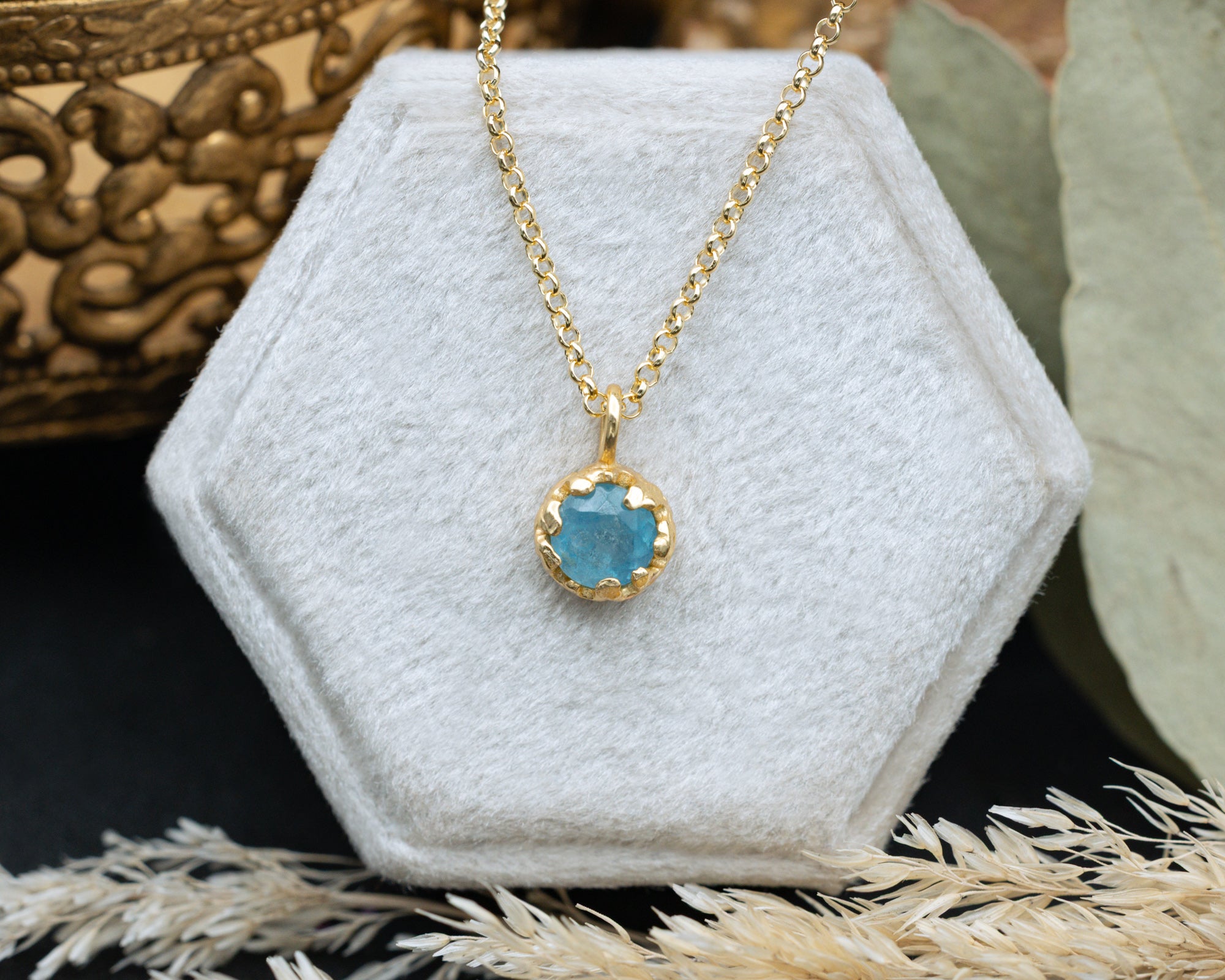 Gold necklace with a blue pendant on a white hexagonal stand.